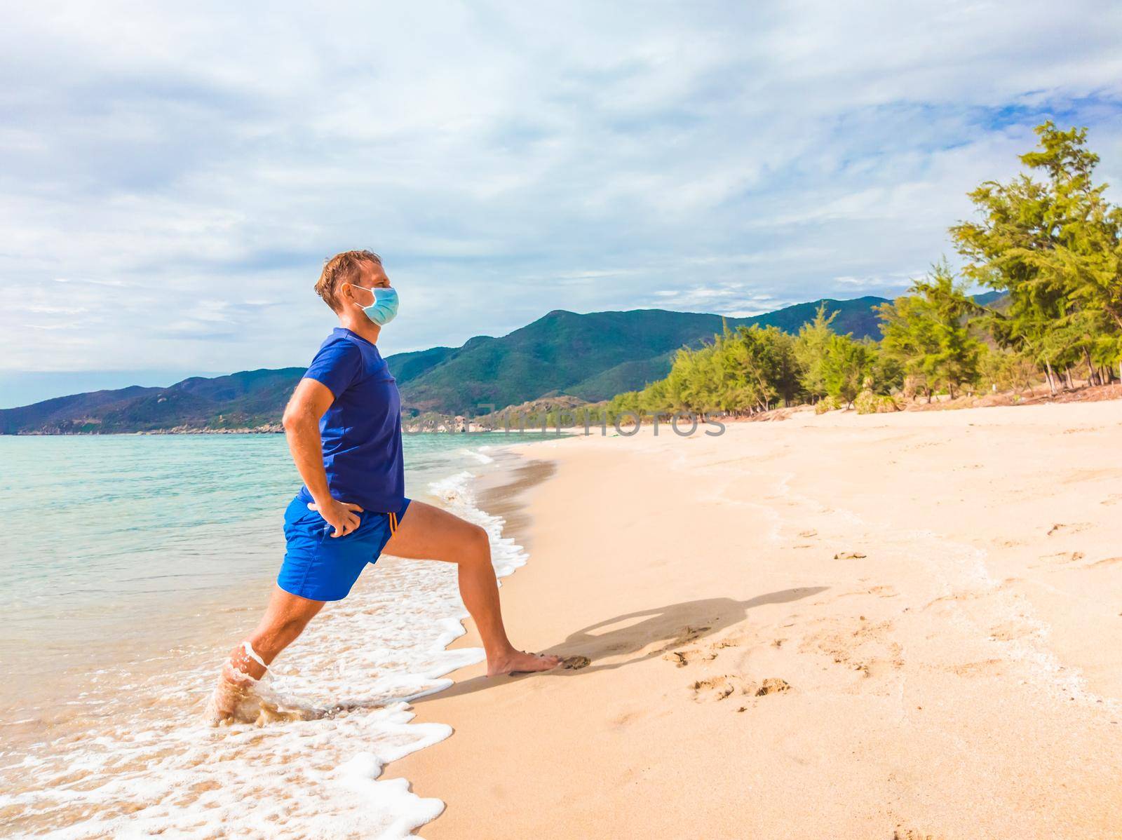 Coronavirus COVID 19. Young beautiful man doing sport performing workouts exercises and running near sea in the morning during quarantine. Active life in surgical sterilizing face mask protection.