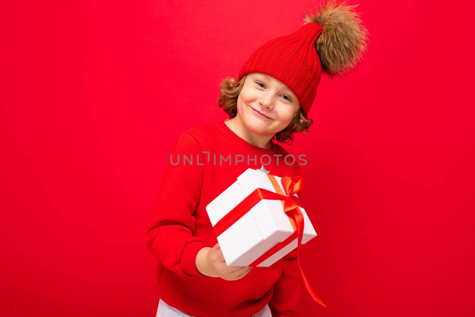 a cool boy with curls on a red wall background in a sweater with a Christmas tree holding a gift box in his hands.