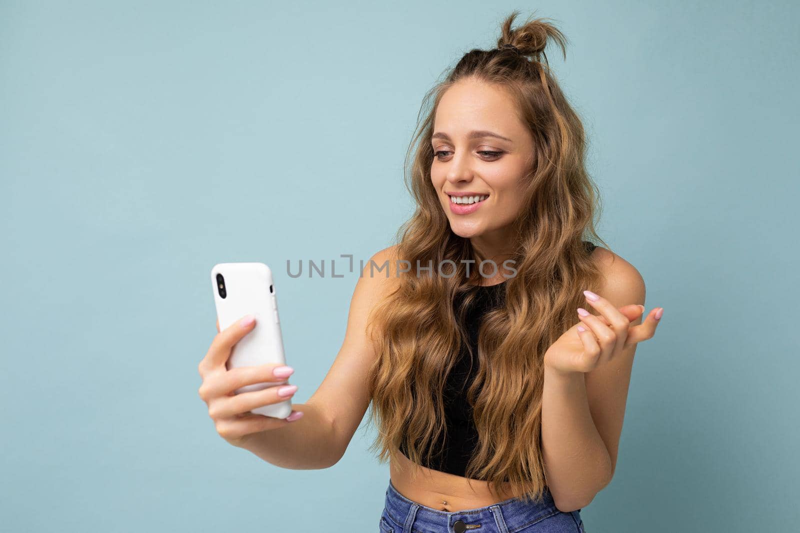 Photo shot of attractive positive good looking young woman wearing casual stylish outfit poising isolated on background with empty space holding in hand and using mobile phone messaging sms looking at smartphone display screen.