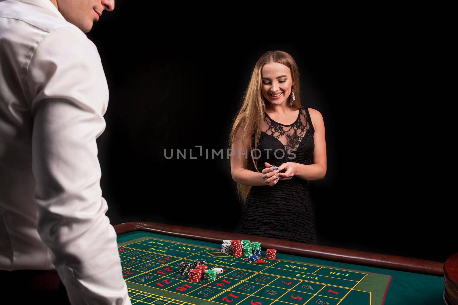 A close-up on the back of the croupier in a white shirt, image of green casino table with roulette and chips, a rich woman betting of gambling in the background. Casino. Gambling. Roulette. Betting