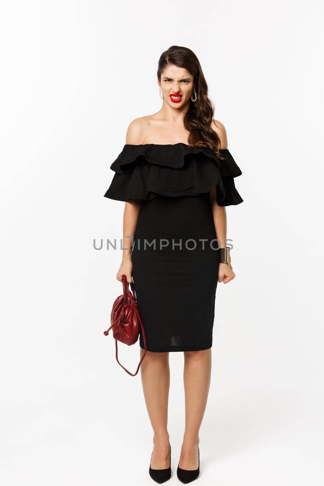 Beauty and fashion concept. Full length of angry woman in black party dress and high heels, express disdain and grimacing at camera, mad at person, white background.