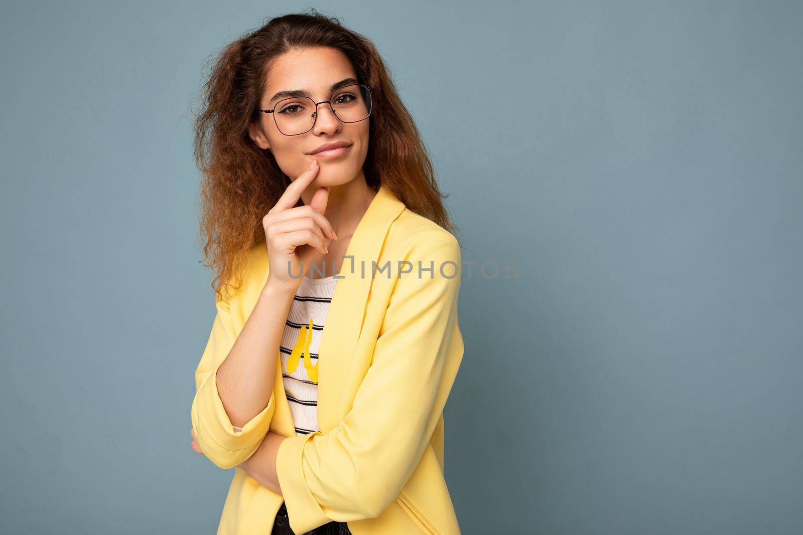 Photo of young positive thinking dreaming beautiful brunette curly woman with sincere emotions wearing casual yellow jacket and optical glasses isolated on blue background with copy space.