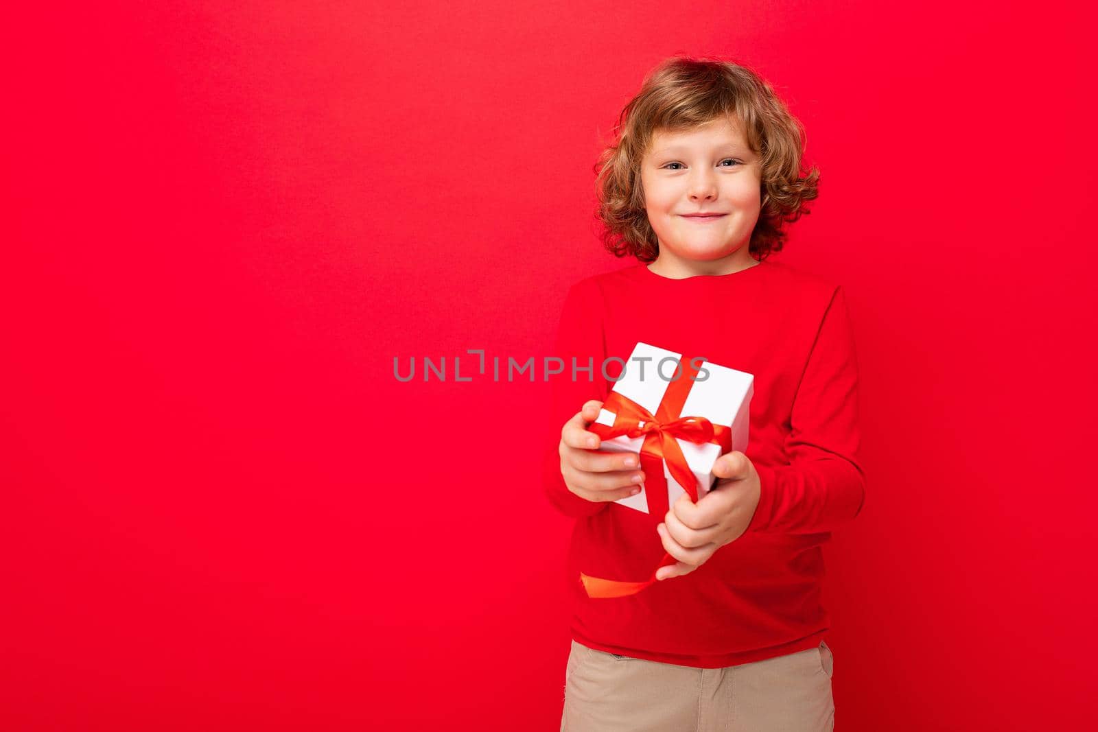 Photo shot of joyful smiling blonde curly boy isolated over red background wall wearing red sweater holding gift box and looking at camera. Copy space