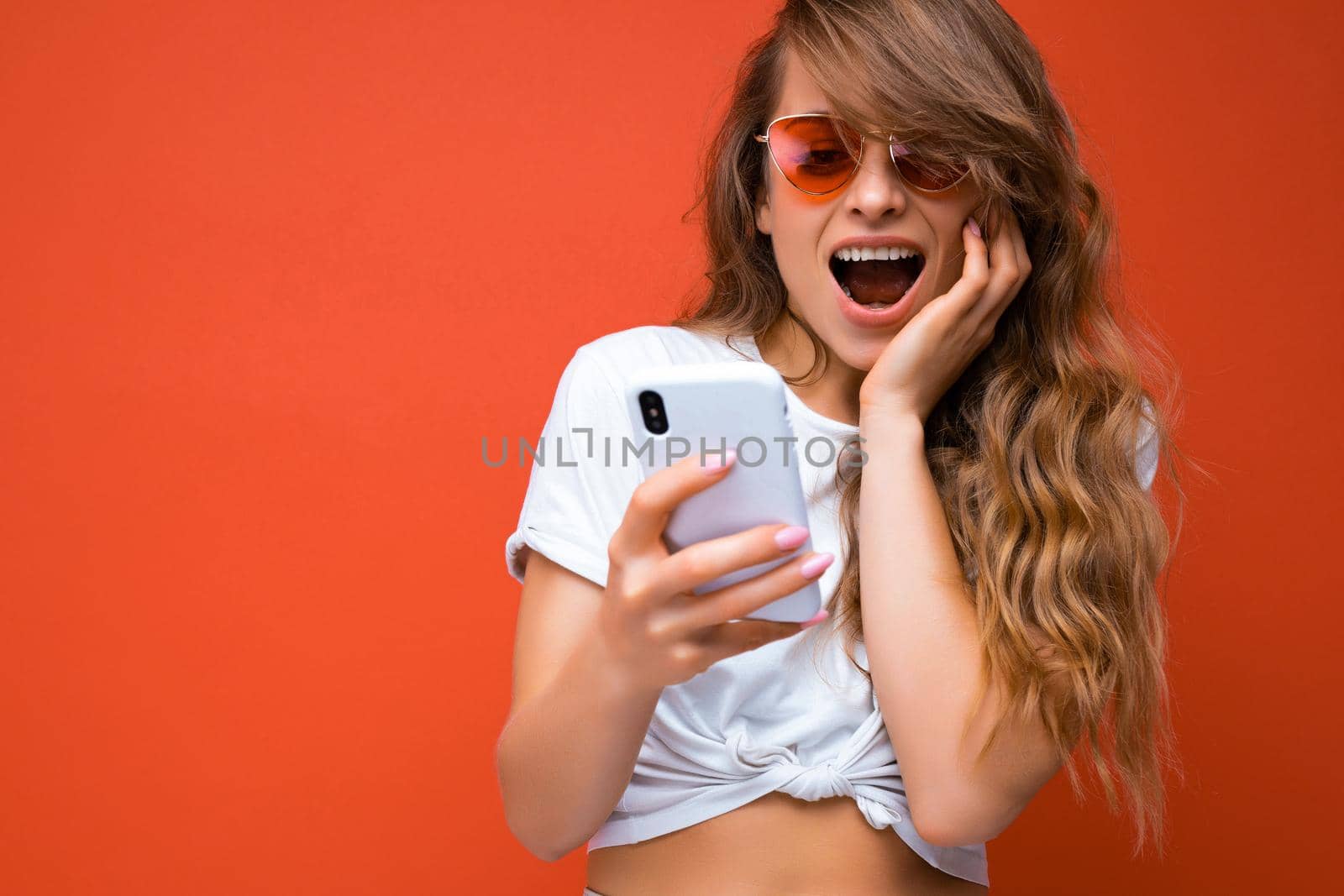 amazed beautiful young woman holding mobile phone wearing sunglasses everyday stylish outfit isolated over wall background looking at device screen and reading news in the internet. Copy space
