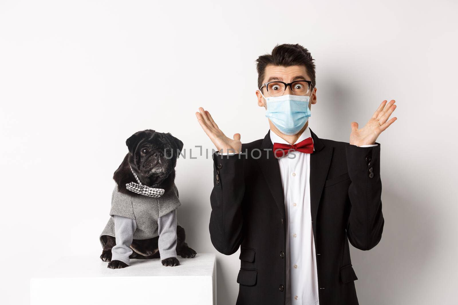 Coronavirus, pets and celebration concept. Amazed young man in face mask and suit staring at camera surprised, cute black dog sitting near owner in party outfit, white background by Benzoix