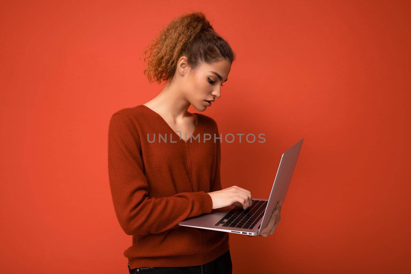 Close-up portrait of beautiful dark blond woman holding laptop computer looking at netbook monitor typing on keyboard wearing red sweater isolated over red wall background.