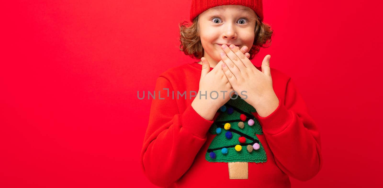 cool teenager in a red Christmas sweater fooling around against the background of a red wall, a warm hat and a sweater with a Christmas tree by TRMK