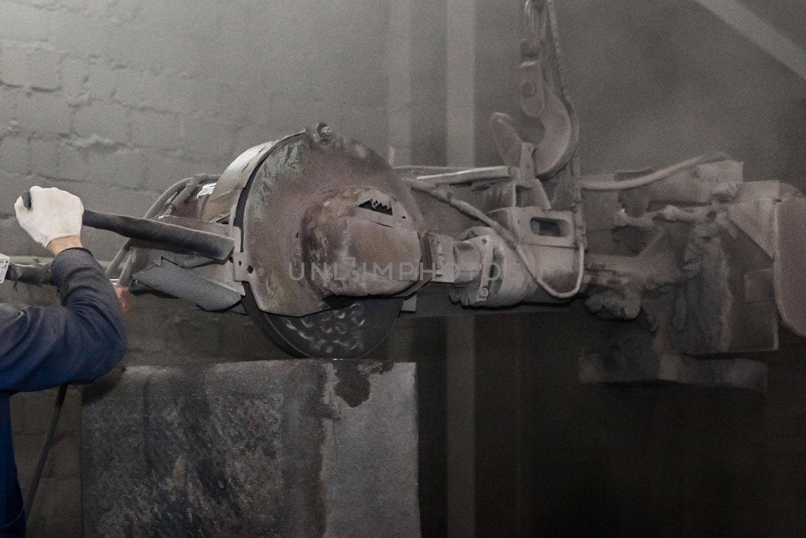 The worker's hands hold and control heavy equipment for grinding and cleaning cast iron tubing in the dirty shop of the industrial plant.