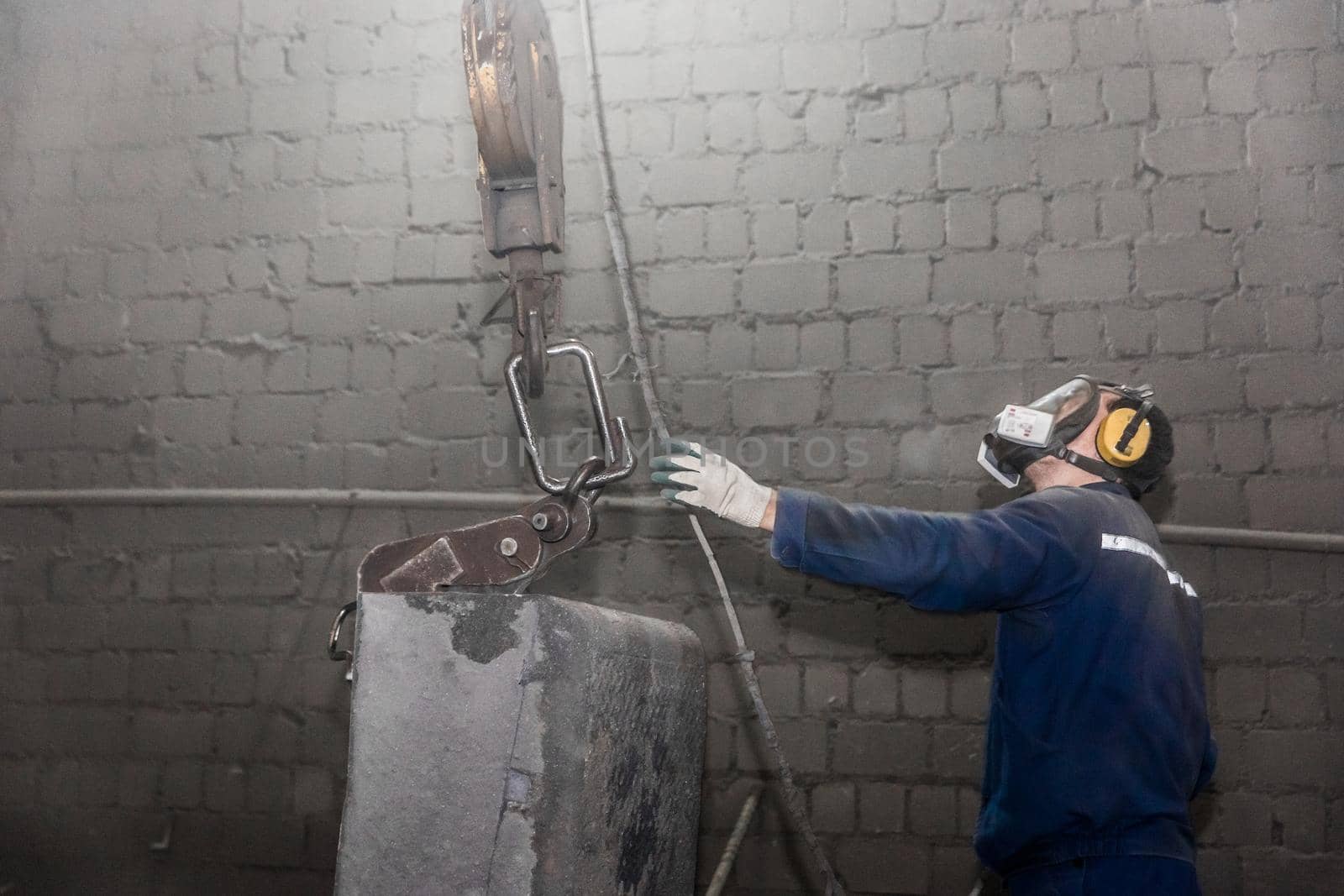 A working man in overalls and a respirator takes a hook with his hand lifting mechanism, equipment with cast iron reinforced concrete tubing in the workshop of an industrial plant.