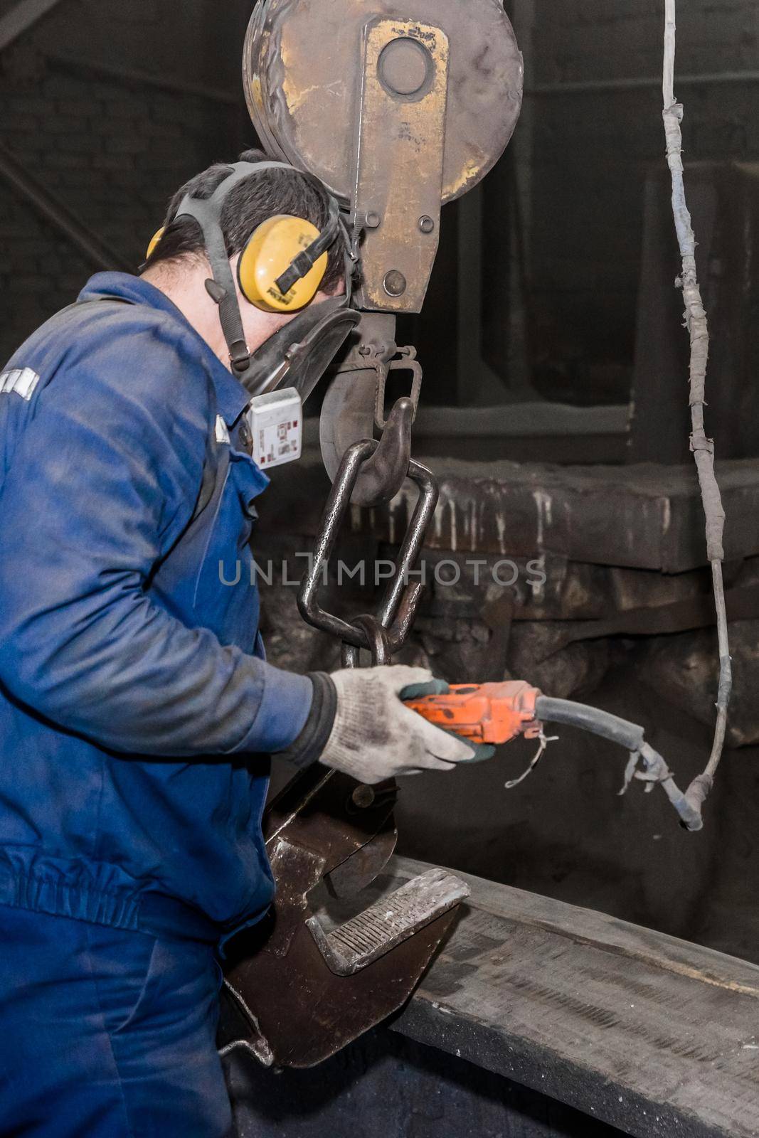 A working man in overalls and a respirator holds in his hand the control panel of the lifting mechanism in front of the cast iron concrete tube, in the dirty workshop of the industrial plant.