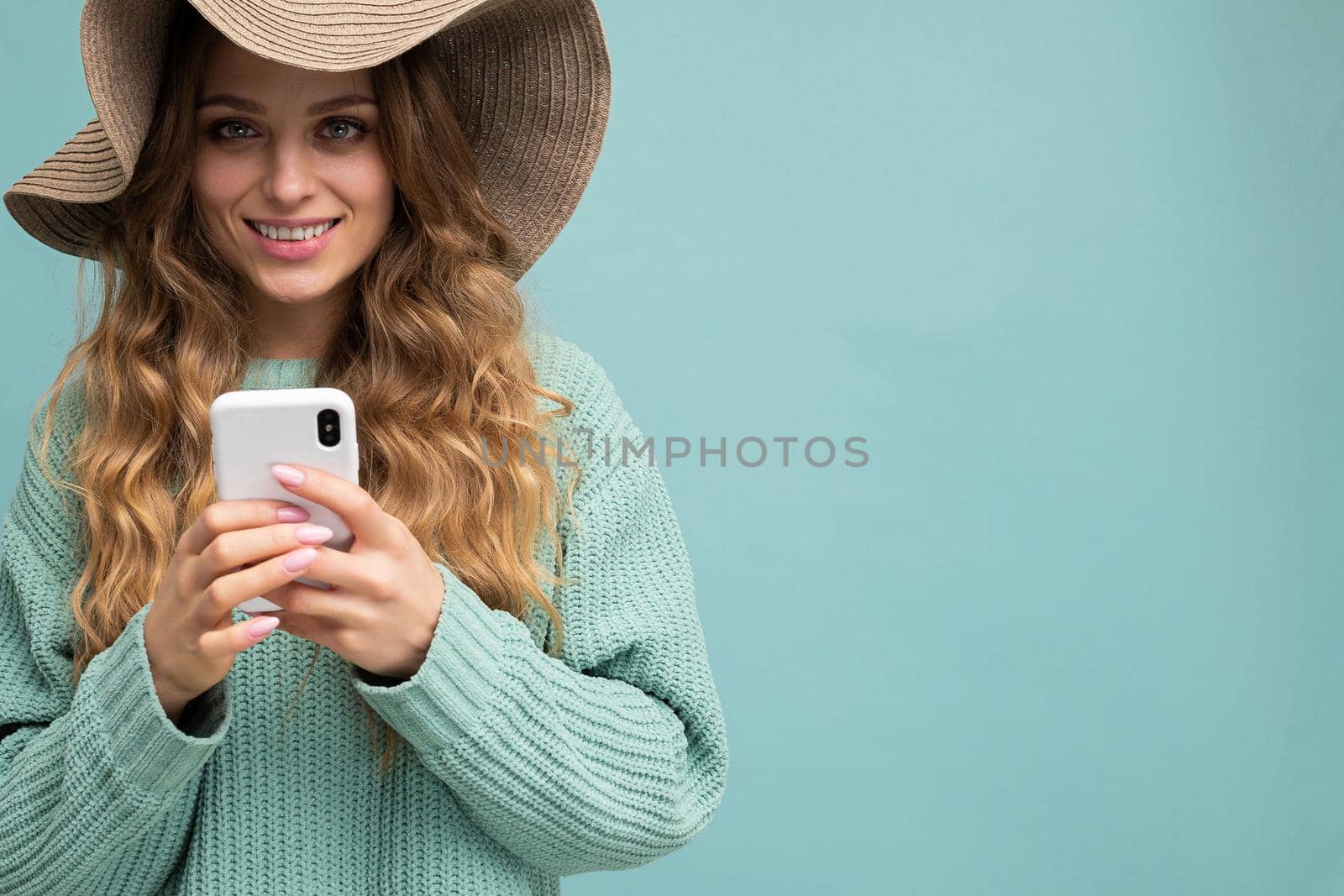 Closeup portrait photo of beautiful young blonde woman wearing blue sweater and hat standing isolated over blue background surfing on the internet via phone looking at camera. Copy space