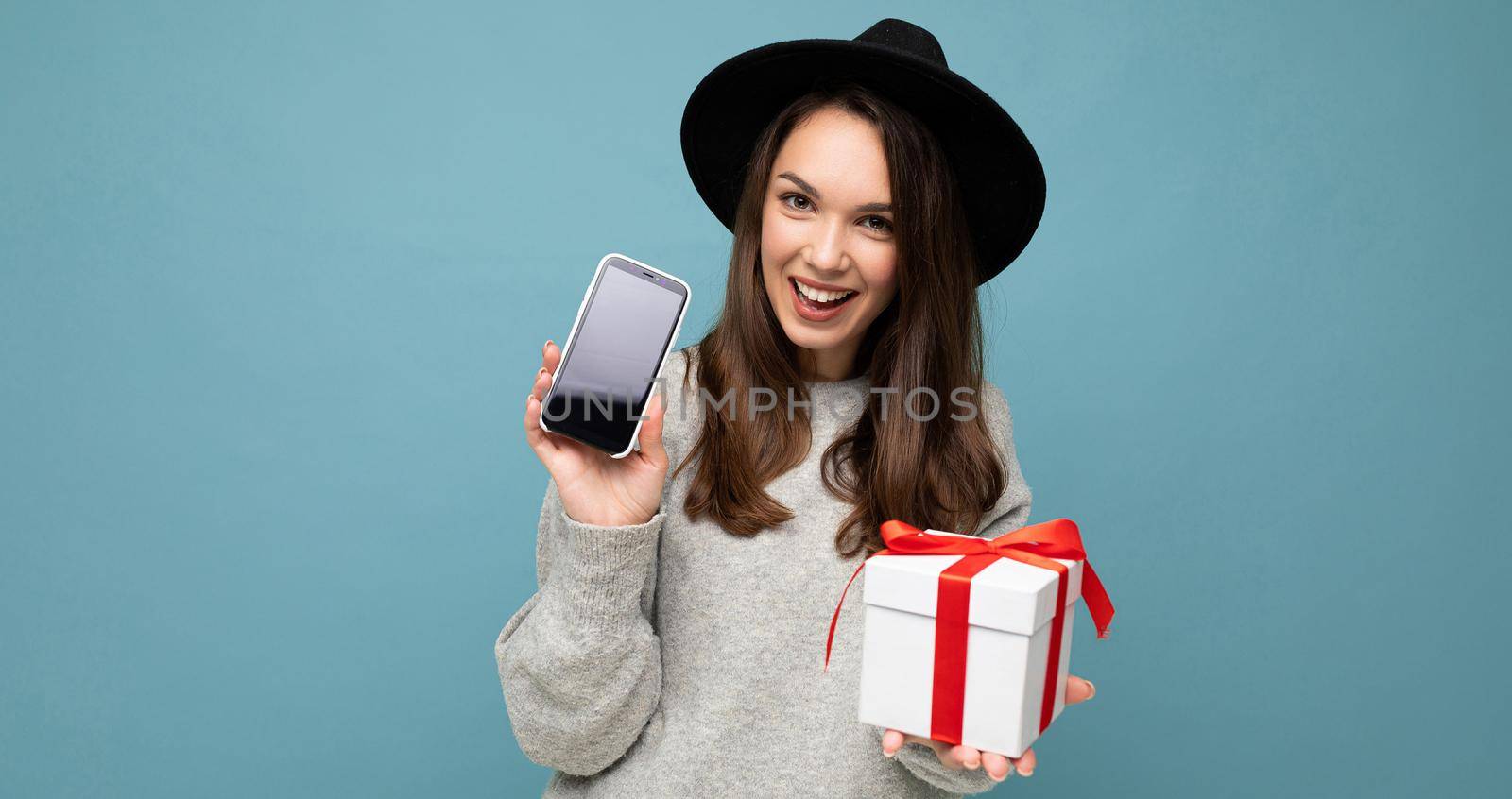 Shot of pretty smiling positive young brunette woman isolated over blue background wall wearing stylish black hat and grey sweater holding gift box showing smartphone screen display for mockup and looking at camera.