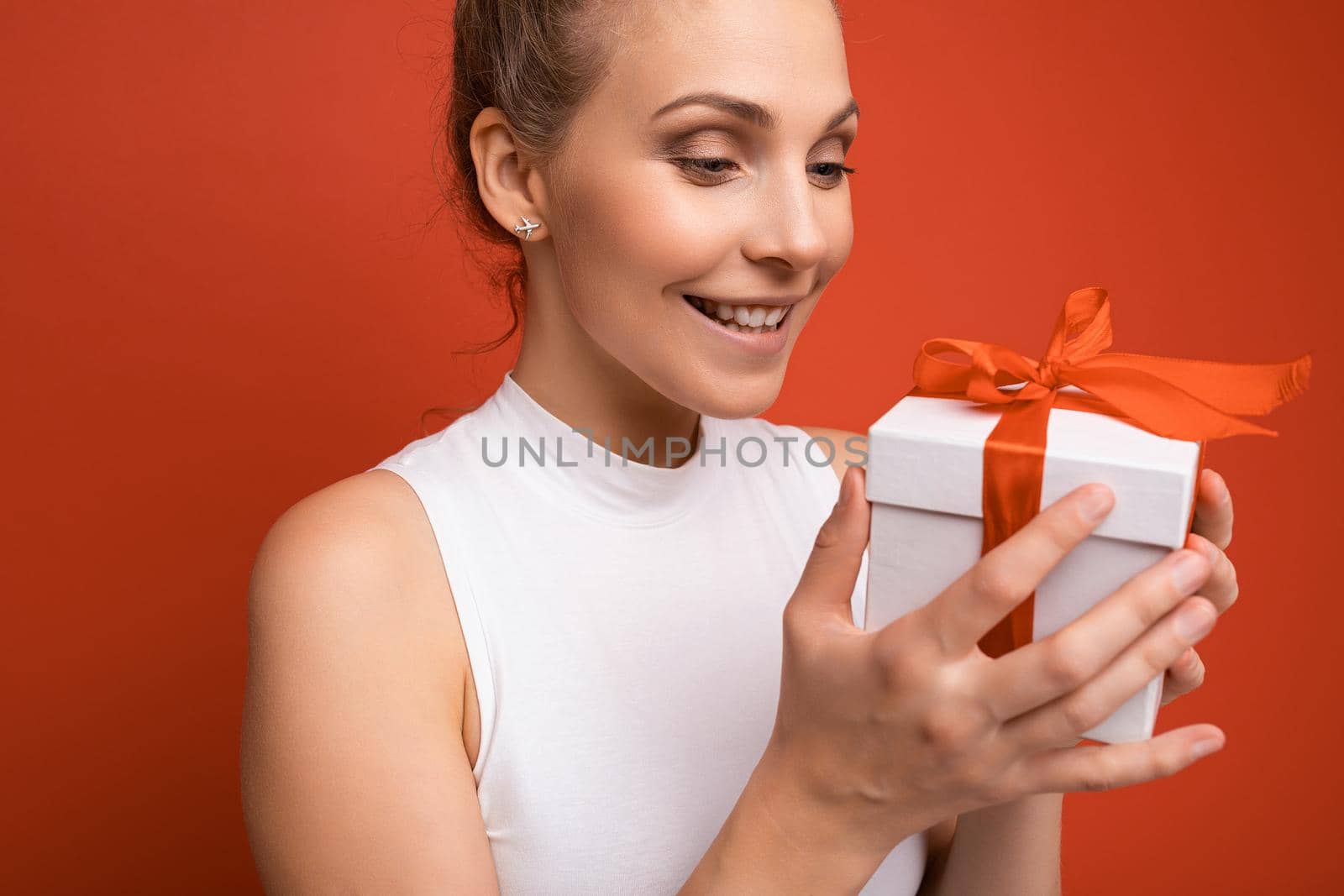 Closeup photo shot of beautiful happy smiling adult blonde woman isolated over red background wall wearing white top holding gift box and looking at white box with red ribbon.