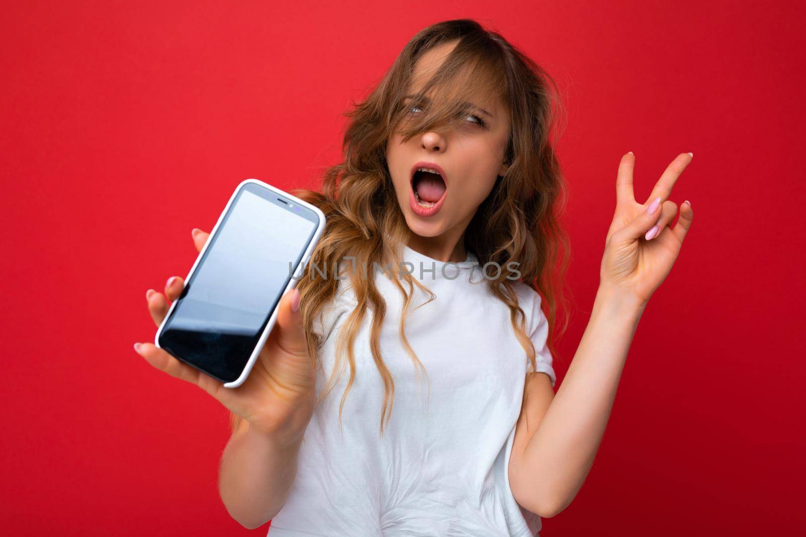 Photo of beautiful amazed astonished young blonde woman good looking wearing white t-shirt standing isolated on red background with copy space holding phone showing smartphone in hand with empty screen display for mockup looking at camera showing peace gesture.