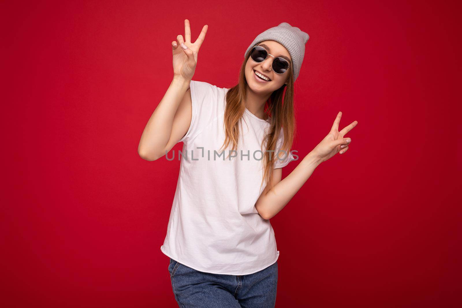 Portrait of young emotional positive happy smiling beautiful dark blonde woman with sincere emotions wearing casual white t-shirt with empty space for mockup gray hat and sunglasses isolated over red background with free space and showing peace gesture.