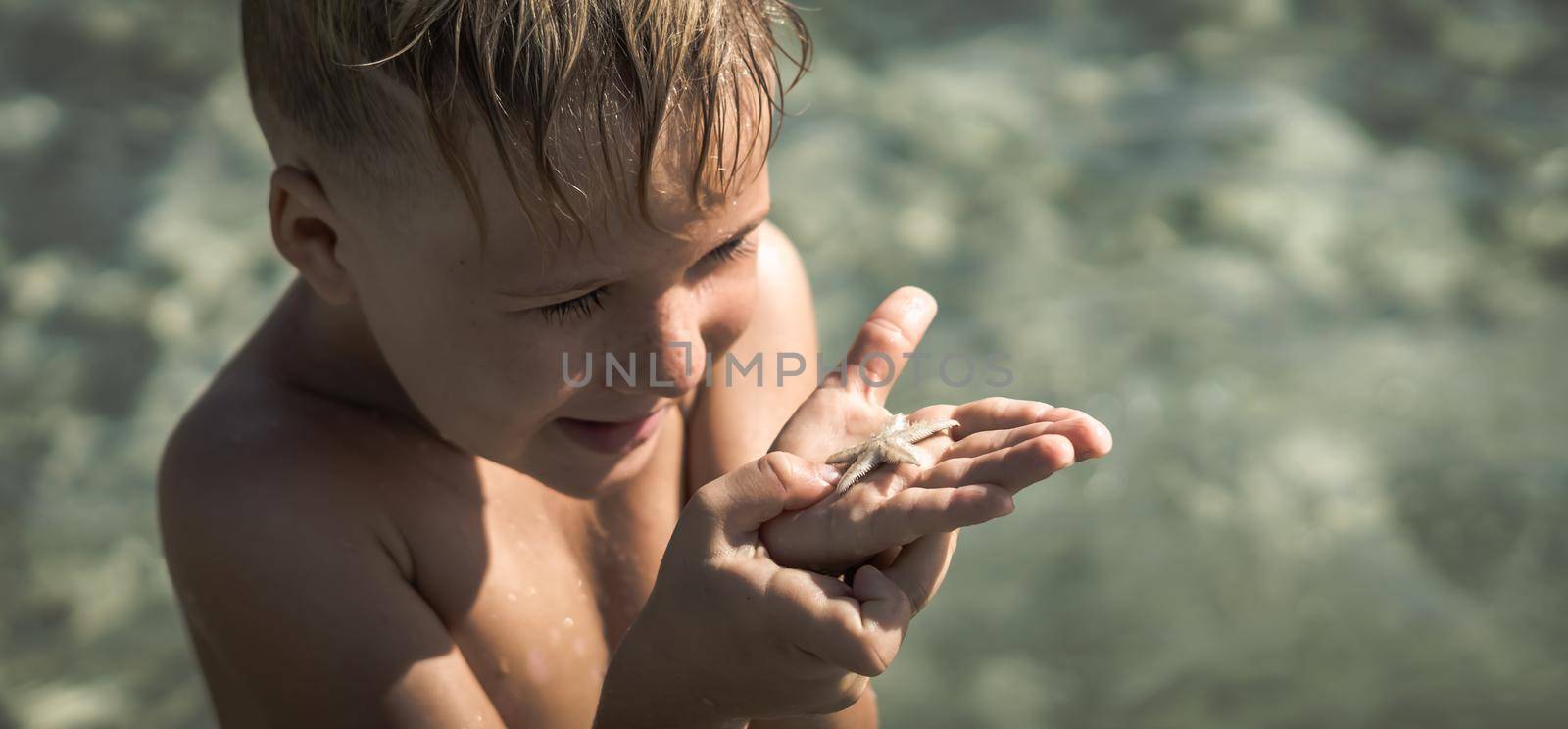 BANNER Copy space. Close side lifestyle portrait boy carefully thoughtful surprise facial expression look at white little starfish on palm. Day sun sea. Childhood learn new interest kid tourism happy.