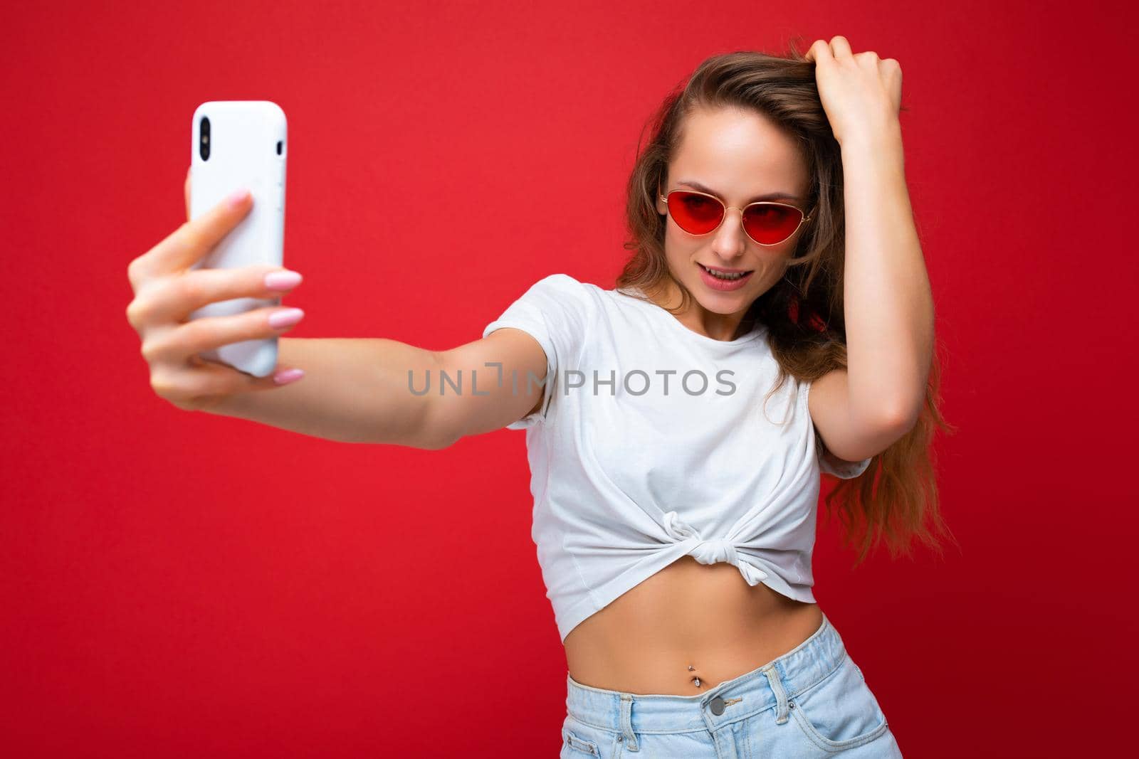 sexy blonde woman holding mobile phone taking selfie photo using smartphone camera wearing sunglasses everyday stylish outfit isolated over colorful wall background looking at device screen.