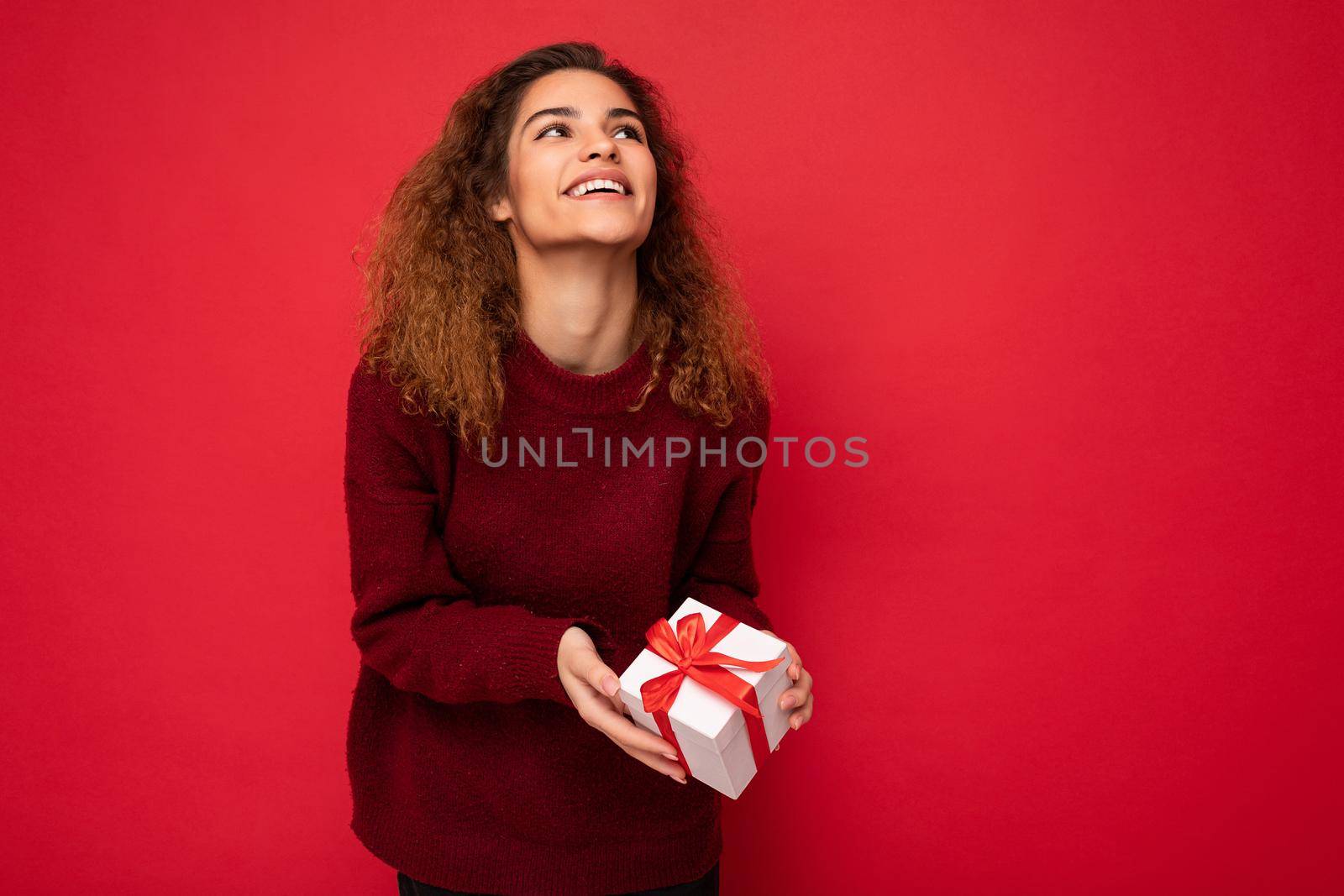 Attractive positivejoyful young brunette curly female person isolated over red background wall wearing red sweater holding gift box looking up. Copy space, mockup