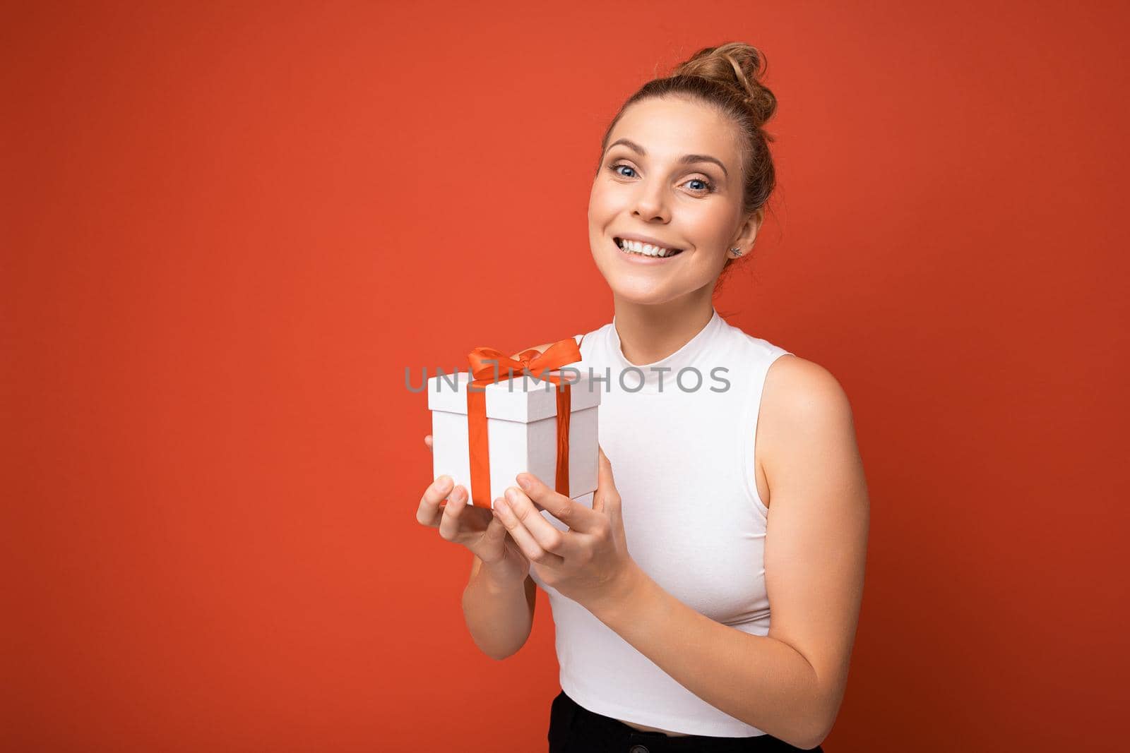 Beautiful happy young blonde woman isolated over colourful background wall wearing stylish casual clothes holding gift box and looking at camera. Copy space