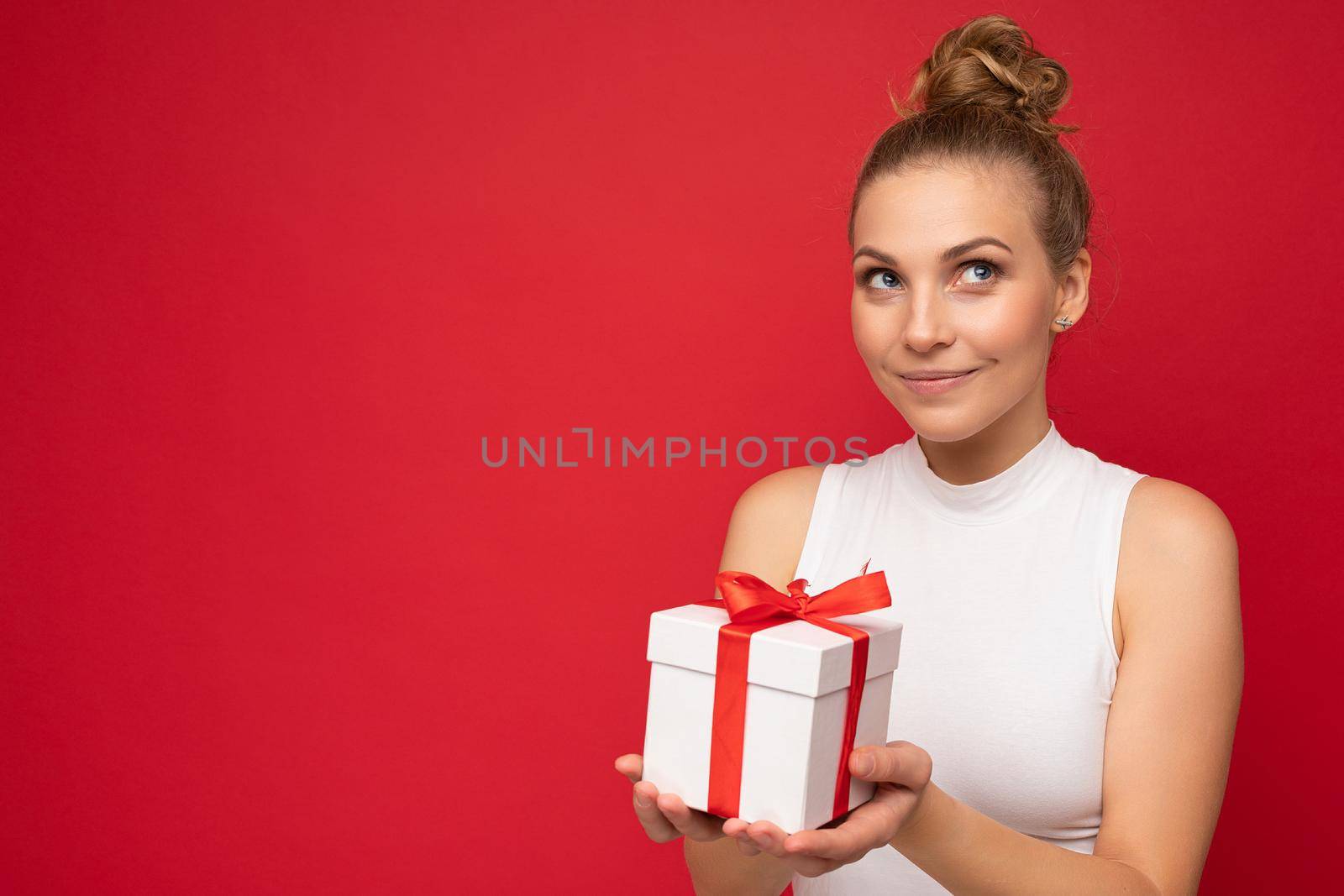 Beautiful happy young blonde woman isolated over colourful background wall wearing stylish casual clothes holding gift box and looking to the side. Copy space, mockup