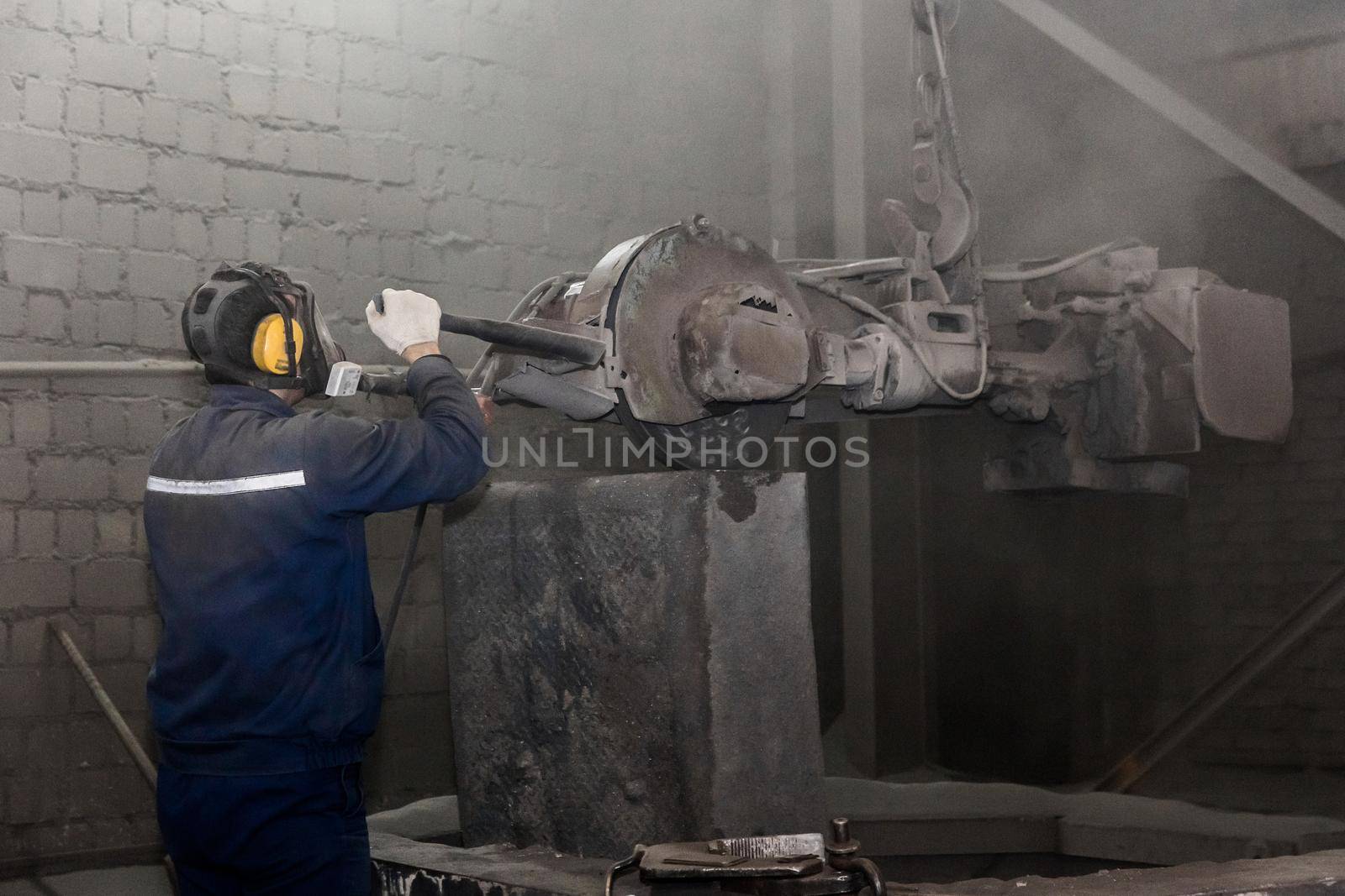 A man in work clothes, and a respirator drives heavy equipment with grinding stone to clean and prepare cast iron concrete tubing in a dirty shop of an industrial plant.