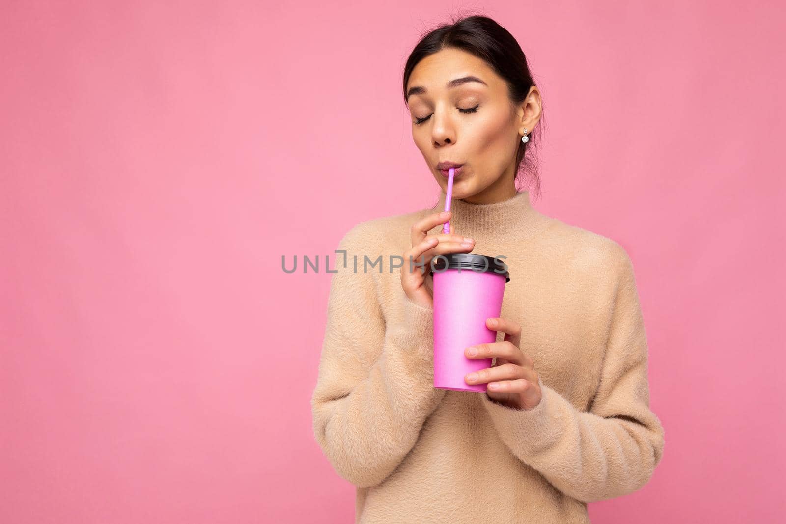 Charming young happy brunette woman wearing stylish clothes isolated over colourful background wall holding paper cup for mockup drinking milkshake enjoying with close eyes. free space