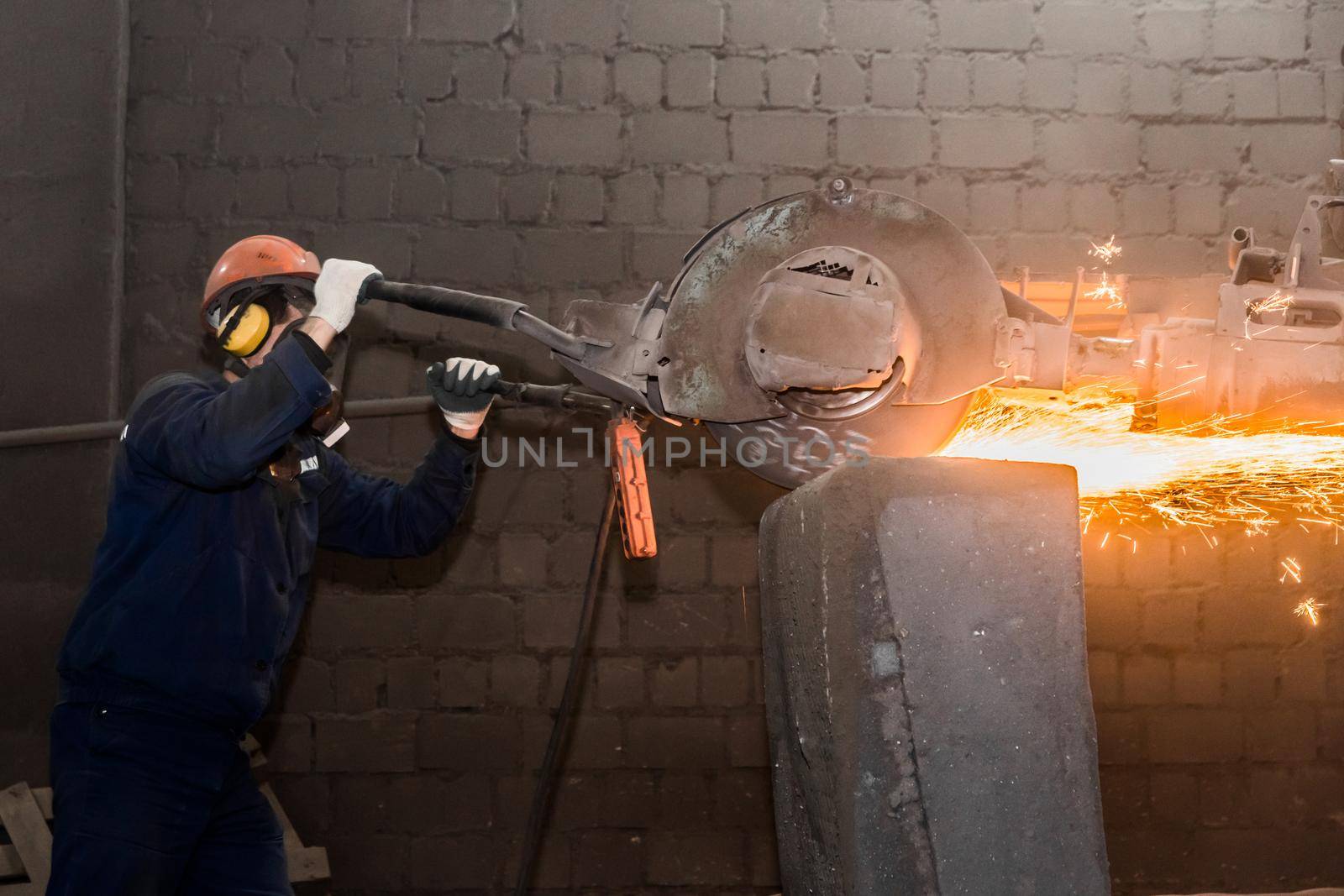 A male worker in a protective helmet, respirator, overalls manages heavy grinding equipment for cast iron concrete tubing with flying sparks in the workshop of an industrial plant.