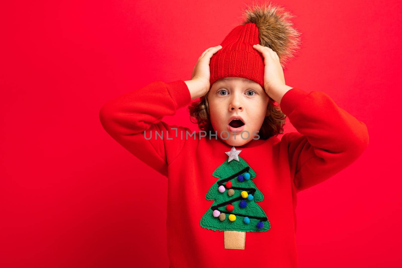 cool teenager in a red Christmas sweater fooling around against the background of a red wall, a warm hat and a sweater with a Christmas tree.