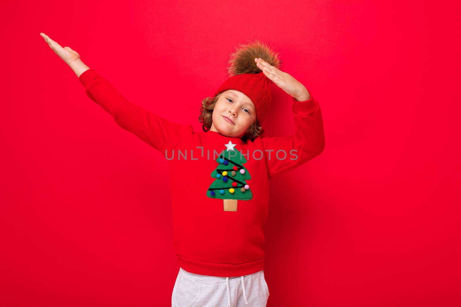cute blond boy in warm hat and christmas sweater on red background with smile on his face.