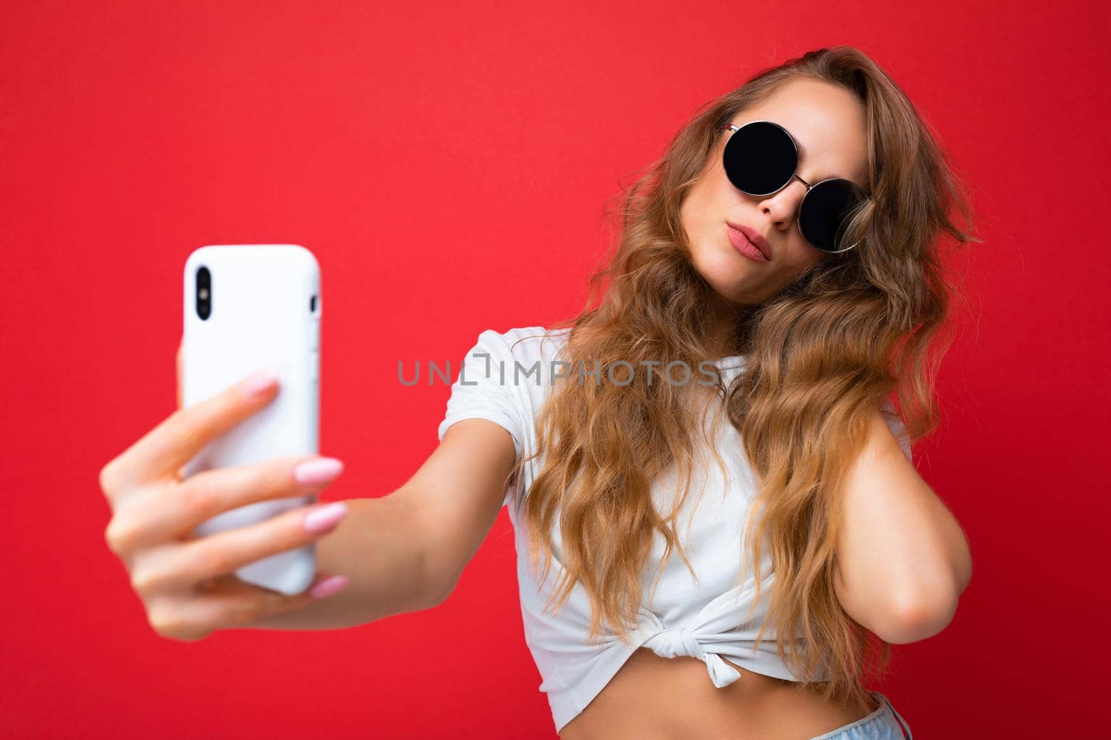 Closeup photo of amazing beautiful young blonde woman holding mobile phone taking selfie photo using smartphone camera wearing sunglasses everyday stylish outfit isolated over colorful wall background looking at device screen.