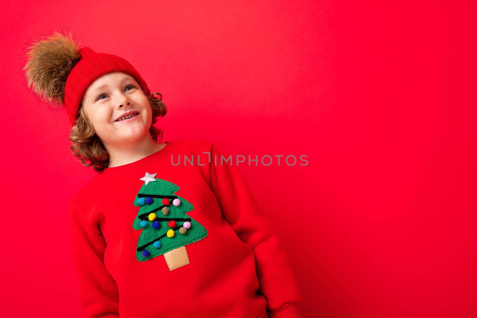 cute blond boy in warm hat and christmas sweater on red background with smile on his face.