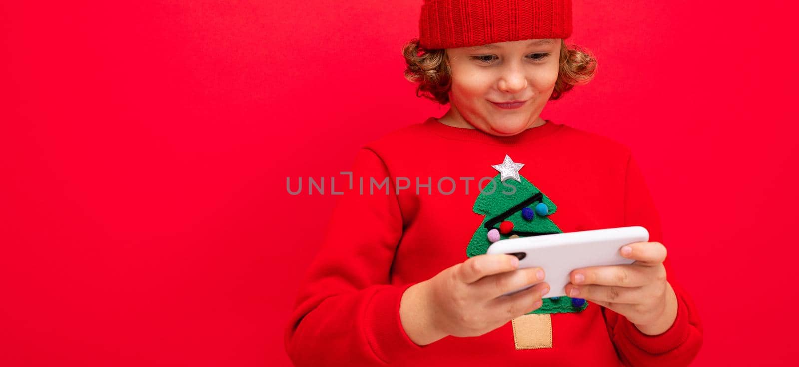 a young man with a smartphone in his hands against the background of a red wall, in a knitted hat and a sweater with a Christmas tree. by TRMK