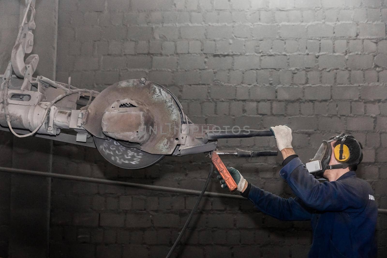 A man in work clothes and a respirator, controls heavy equipment with grinding stone to clean reinforced concrete structures in a dirty shop of an industrial plant by AYDO8