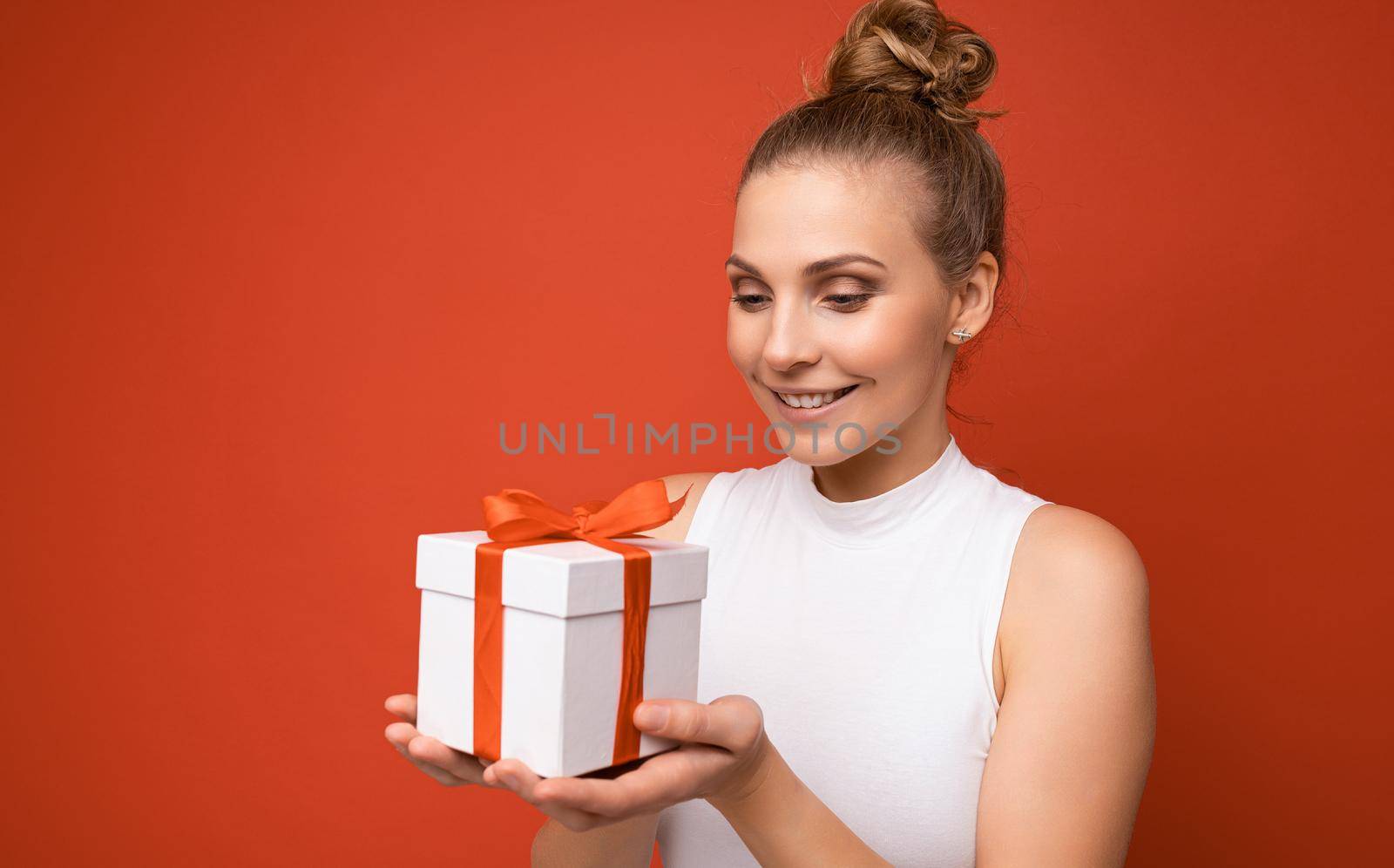 Photo shot of beautiful positive smiling young blonde woman isolated over red background wall wearing white top holding gift box and looking at present.