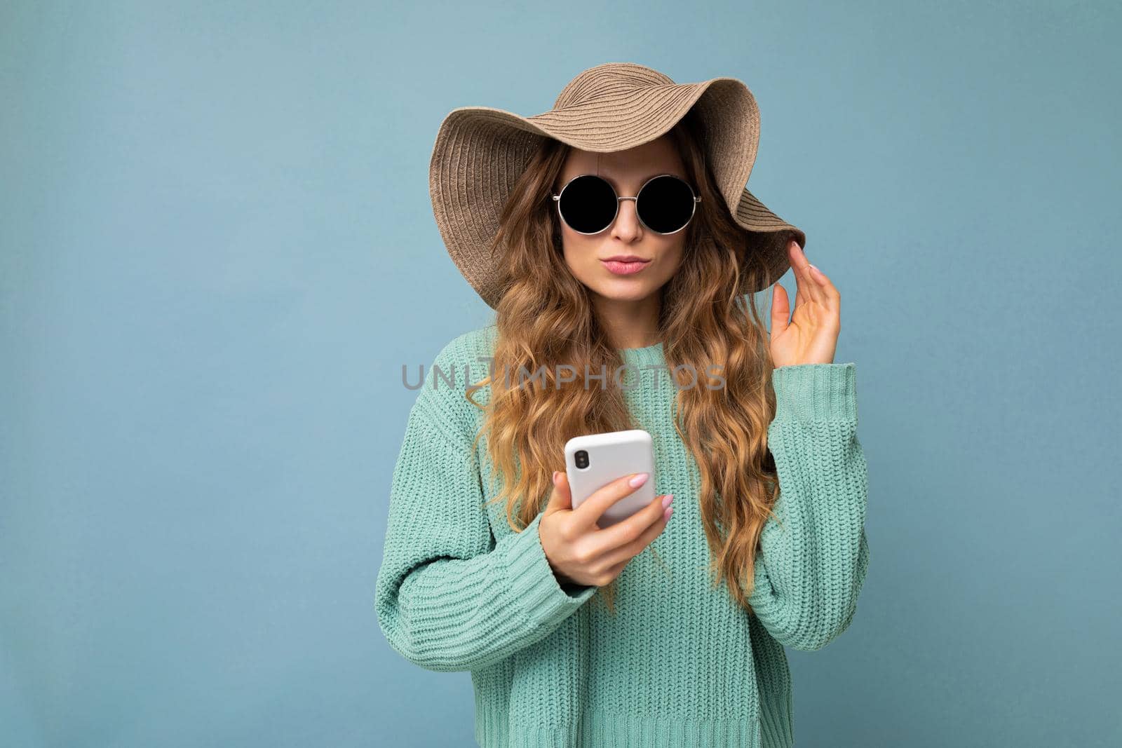 Beautiful young blonde woman wearing blue sweater hat and sunglasses standing isolated over blue background surfing on the internet via phone looking at gadjet.