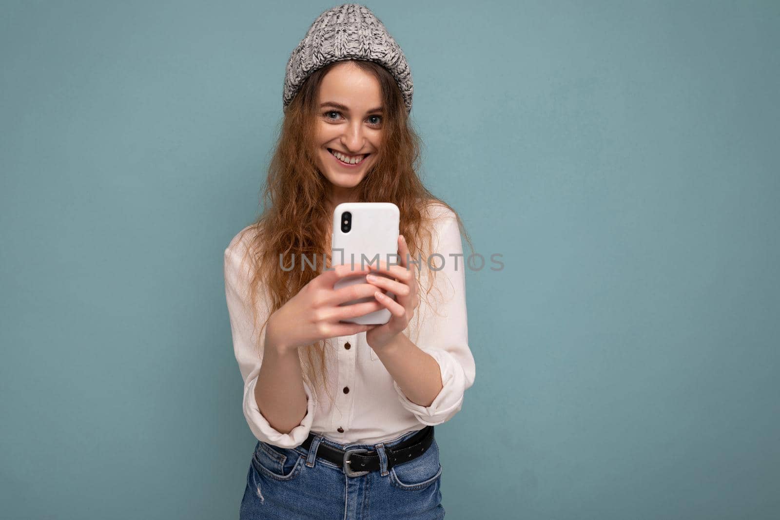 photo of attractive positive young blonde curly woman wearing casual white shirt and grey hat isolated over blue background wall holding phone and using communicating online looking at camera.