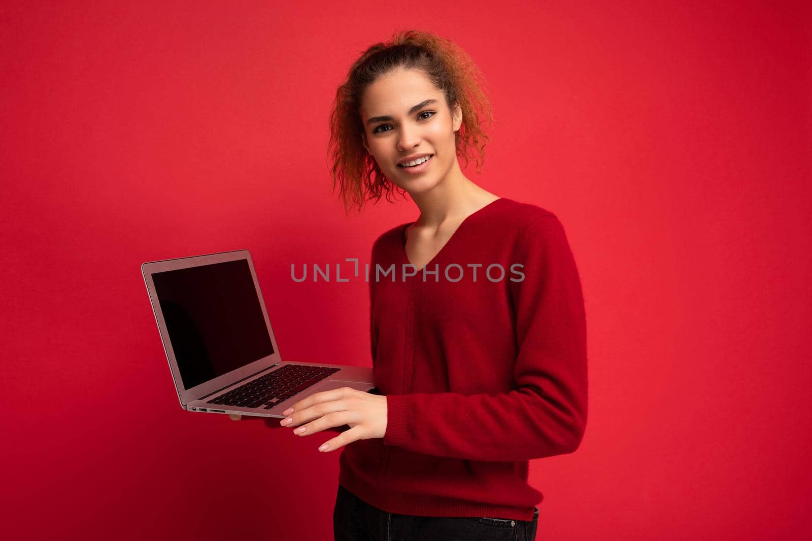 Close-up portrait of beautiful asking and surprised dark blond woman holding laptop computer looking at camera typing on keyboard wearing red sweater isolated over red wall background.