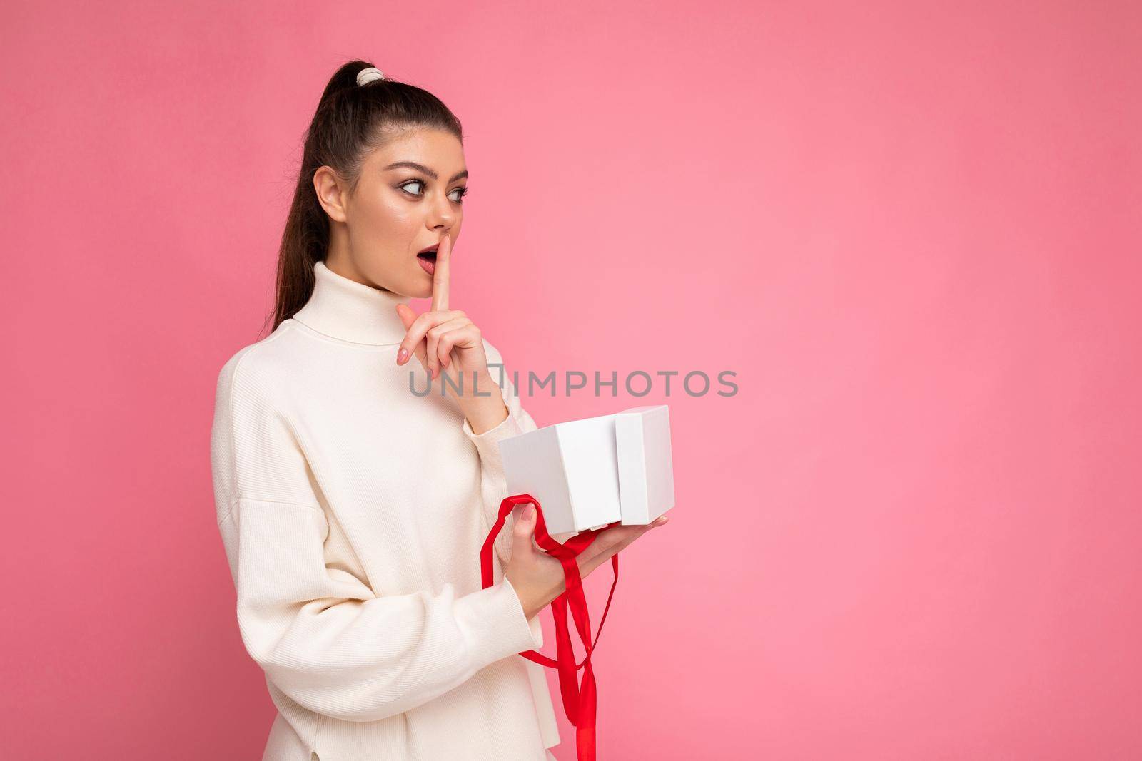 Photo shot of attractive shocked young brunette woman isolated over pink background wall wearing white sweater holding gift box and unboxing present lookingto the side and showing shh gesture. Empty space