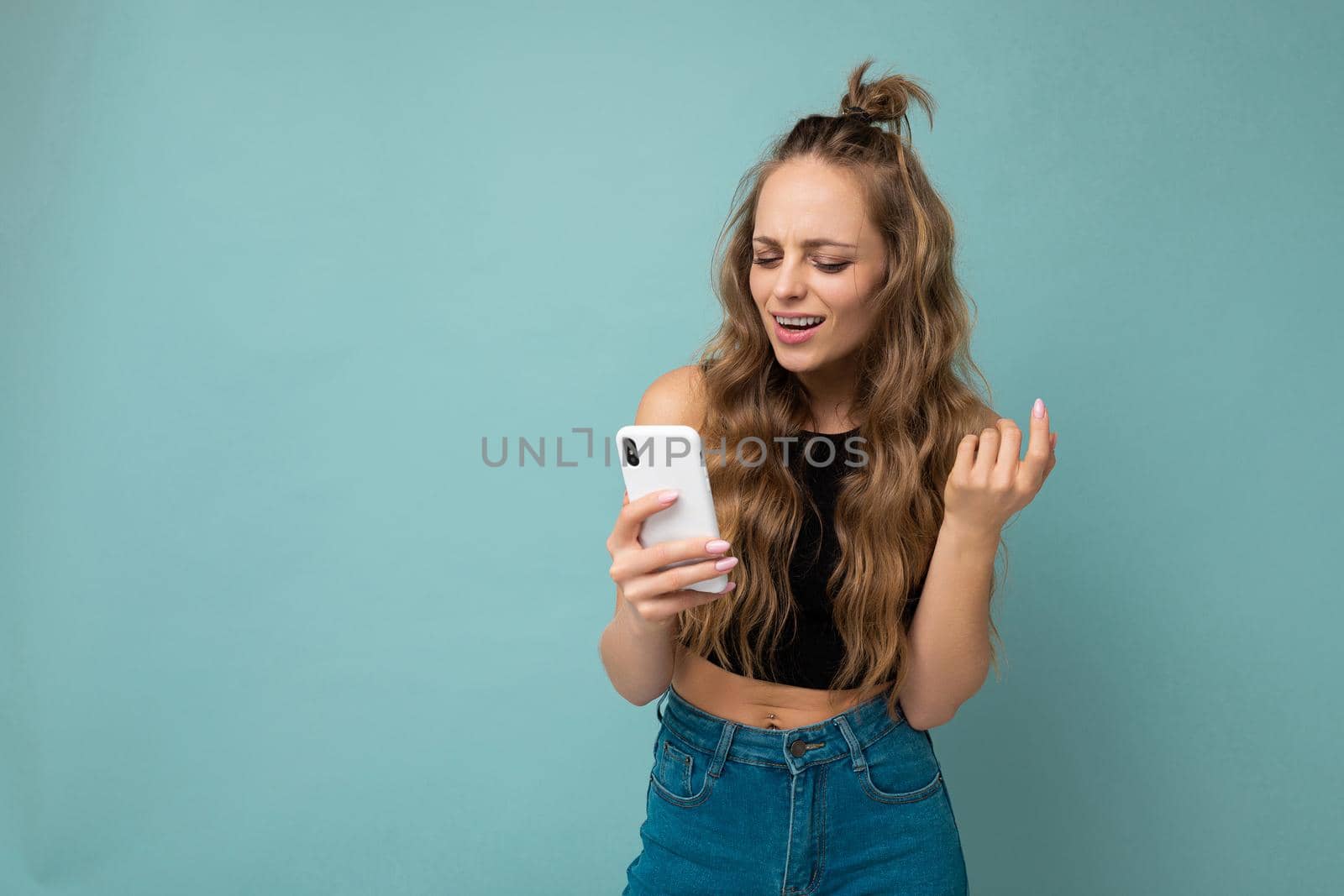 Photo shot of attractive positive good looking young woman wearing casual stylish outfit poising isolated on background with empty space holding in hand and using mobile phone messaging sms looking at smartphone display screen.