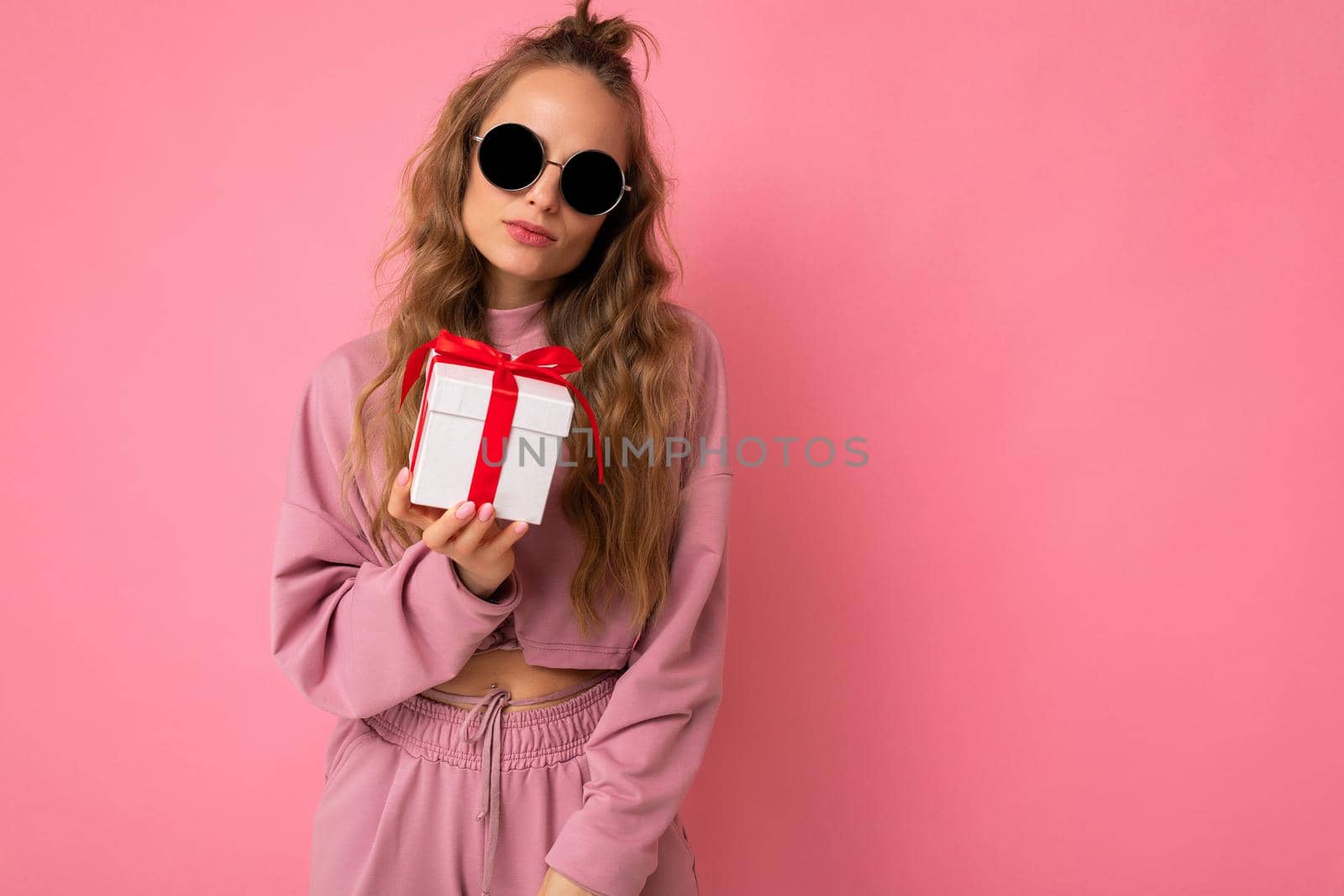 Shot of attractive serious calm young blond curly woman isolated over pink background wall wearing pink sport clothes and modern sunglasses holding gift box looking at camera. Empty space
