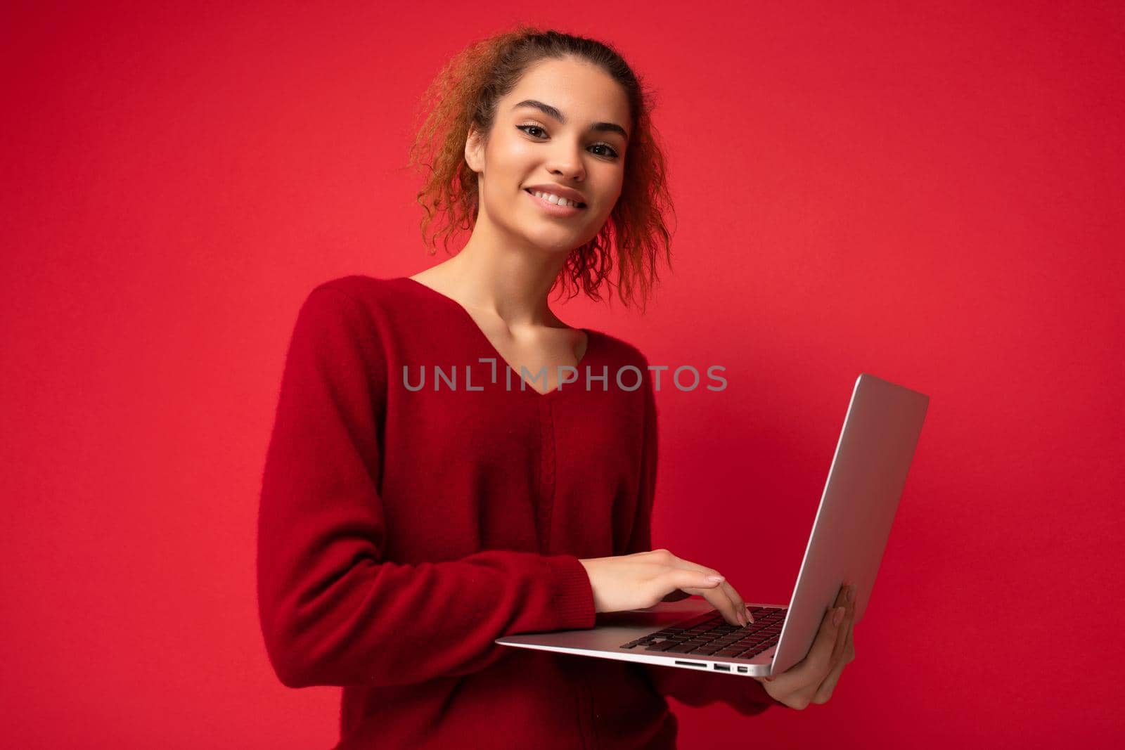 Close-up portrait of beautiful smiling fascinating charming dark blond woman holding laptop computer looking at camera typing on keyboard wearing red sweater isolated over red wall background.