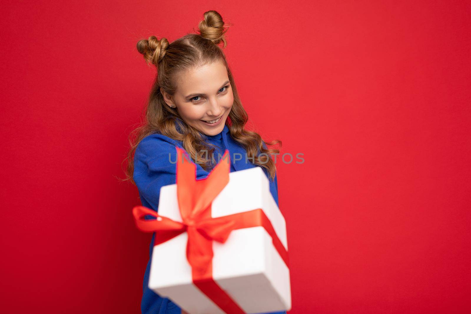 Charming happy funny joyful young blonde woman isolated over red background wall wearing blue trendy hoodie holding gift box and looking at camera.