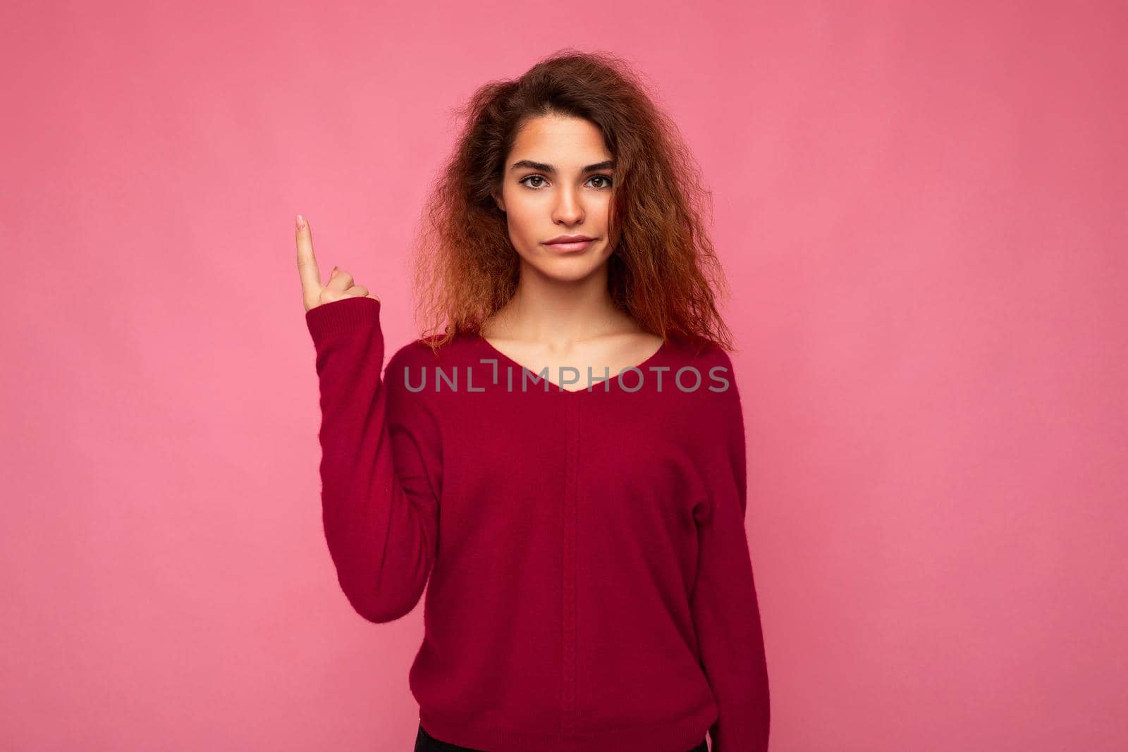 Photo of young serious beautiful brunette wavy woman with sincere emotions wearing casual pink jersey isolated over pink background with copy space and pointing at free space for mockup.