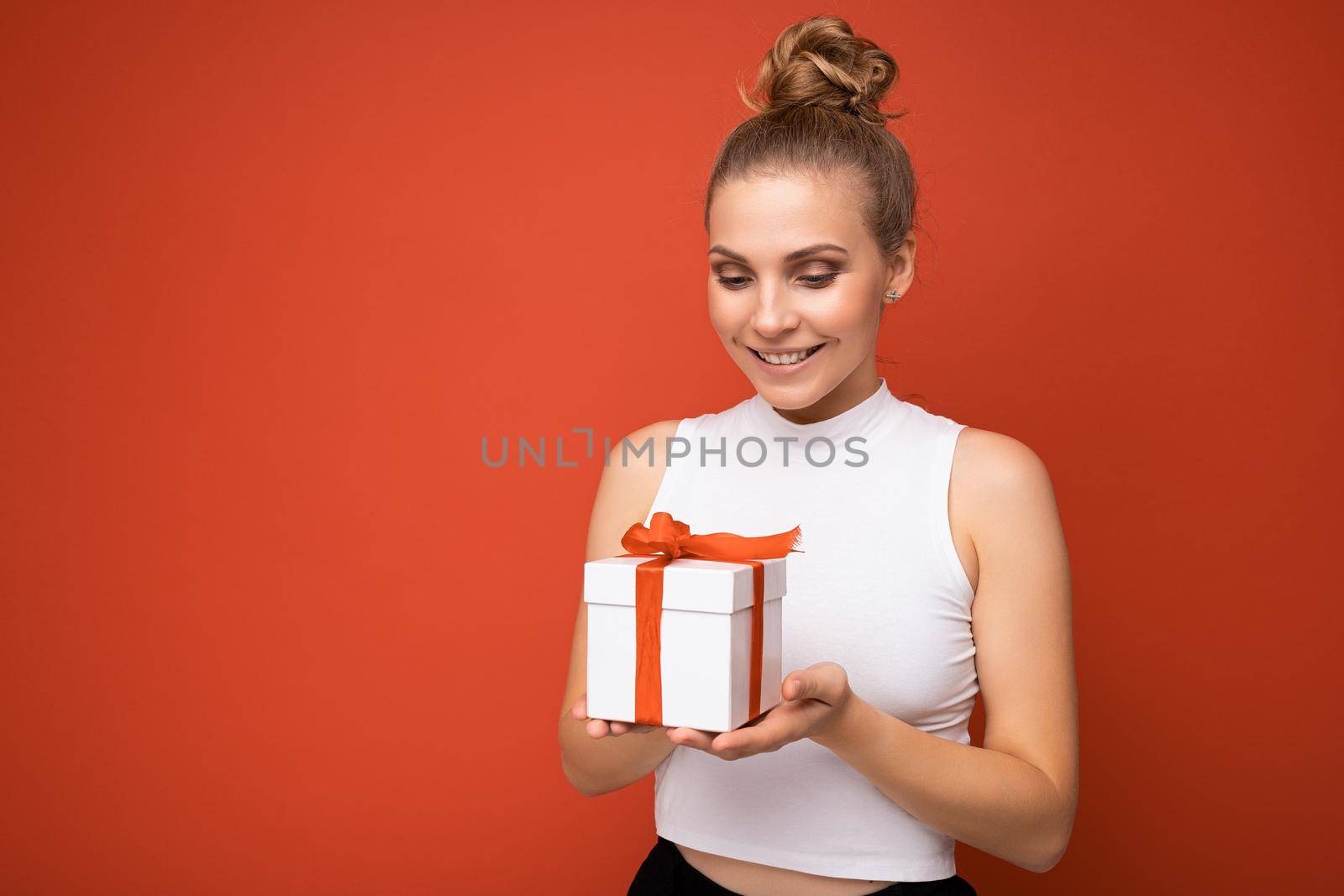 Shot of beautiful positive young blonde woman isolated over red background wall wearing white top holding gift box and looking at present.