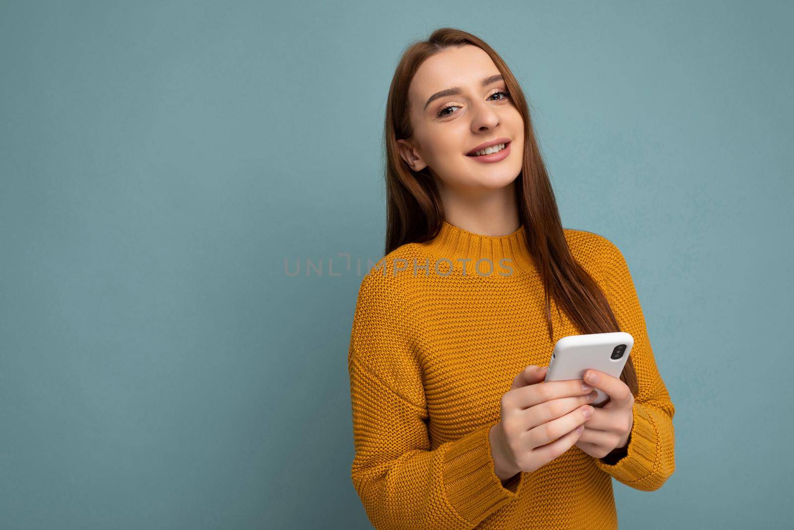 Photo shot of attractive positive good looking young woman wearing casual stylish outfit poising isolated on background with empty space holding in hand and using mobile phone messaging sms looking at camera.