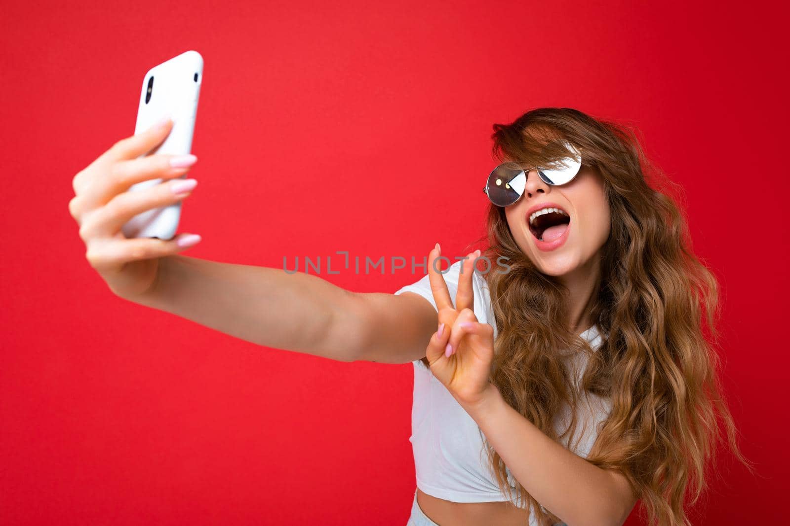 Closeup photo of beautiful young woman holding mobile phone taking selfie photo using smartphone camera wearing sunglasses everyday stylish outfit isolated over colorful wall background looking at device screen and showing peace gesture.