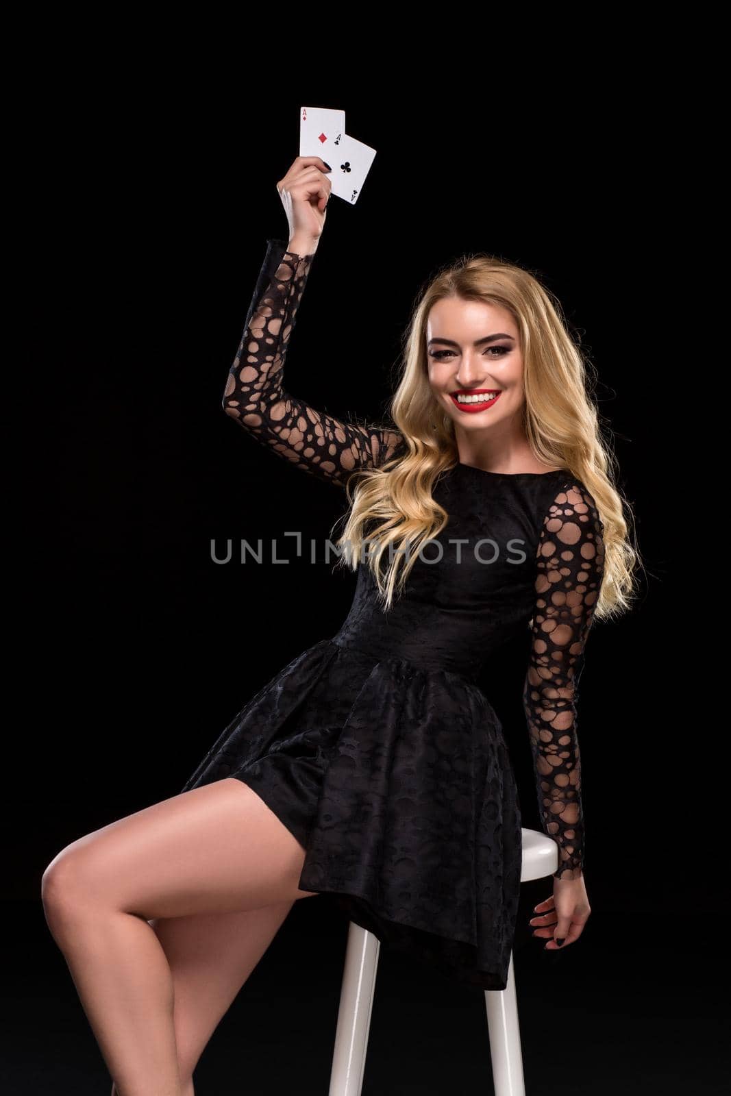 Beautiful confident woman in black dress sitting on chair showing cards looking at camera, isolated on the black background. With copy place Poker. Casino. Roulette Blackjack Spin.