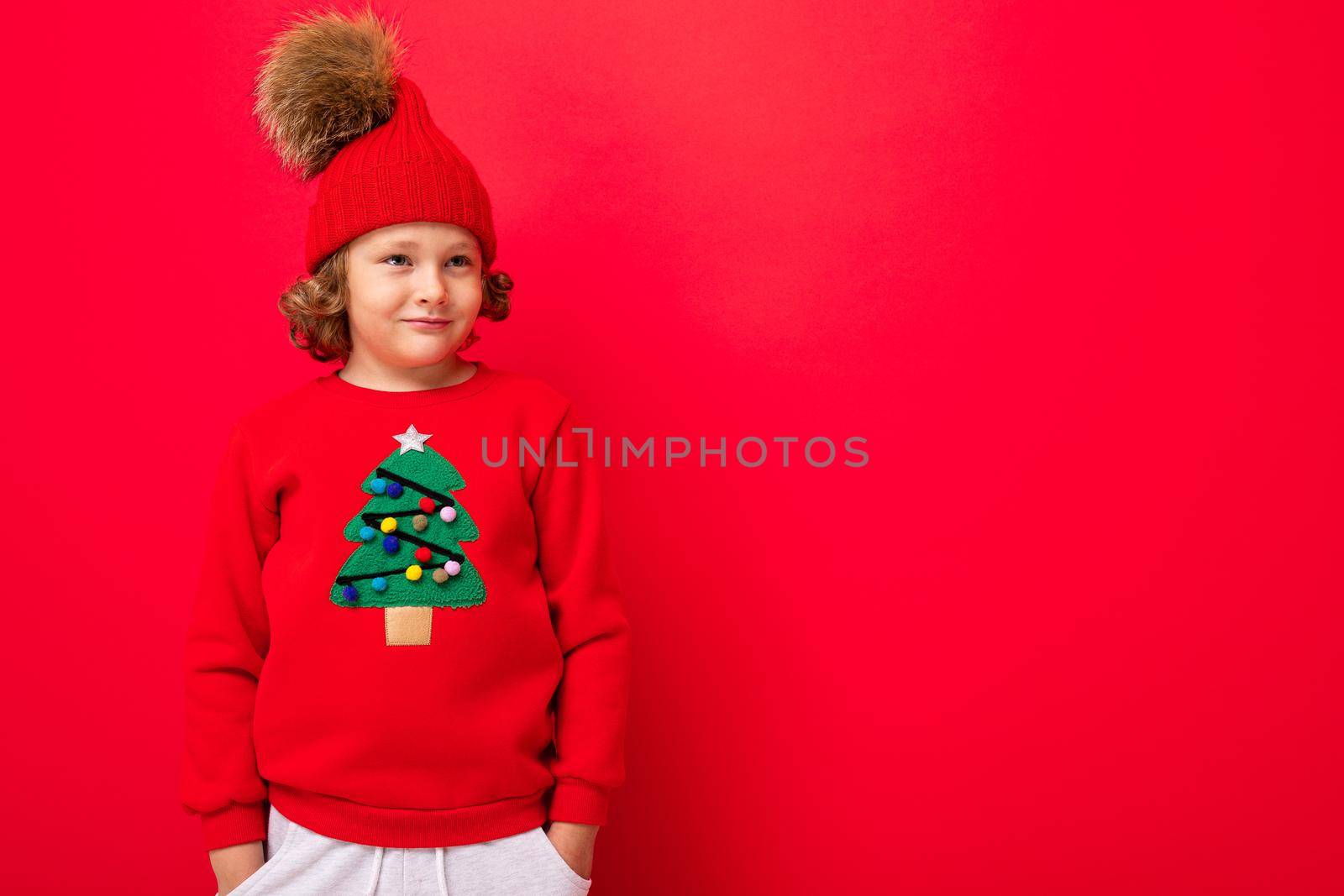 cute blond boy in warm hat and christmas sweater on red background with smile on his face.