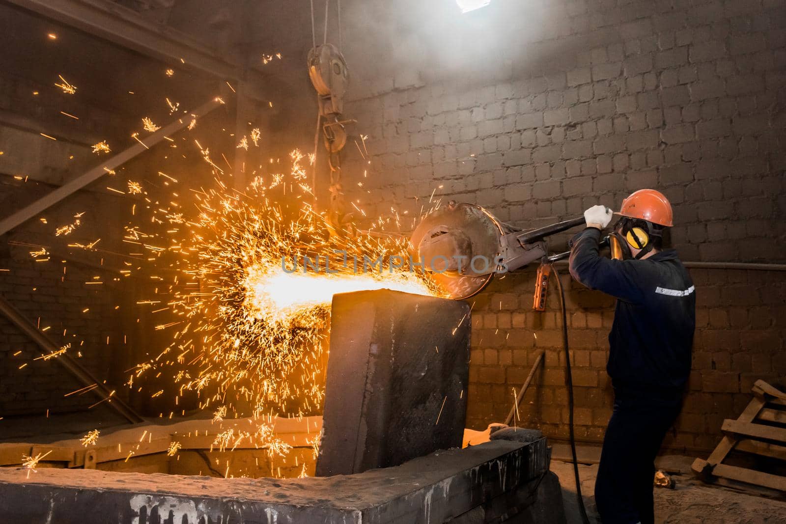 A male worker in a protective helmet, respirator, overalls manages heavy grinding equipment for cast iron concrete tubing with flying sparks in the workshop of an industrial plant by AYDO8