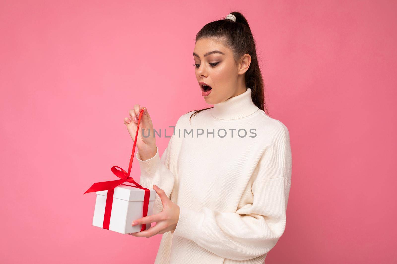 Photo shot of pretty positive surprised young brunet woman isolated over colourful background wall wearing trendy outfit look holding gift box and looking at present box with red ribbon.